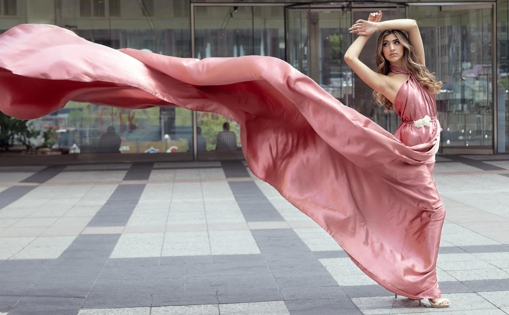 A woman in a flowing pink dress standing outdoors in front of a modern glass building, with her arms raised above her head, looking to the side.