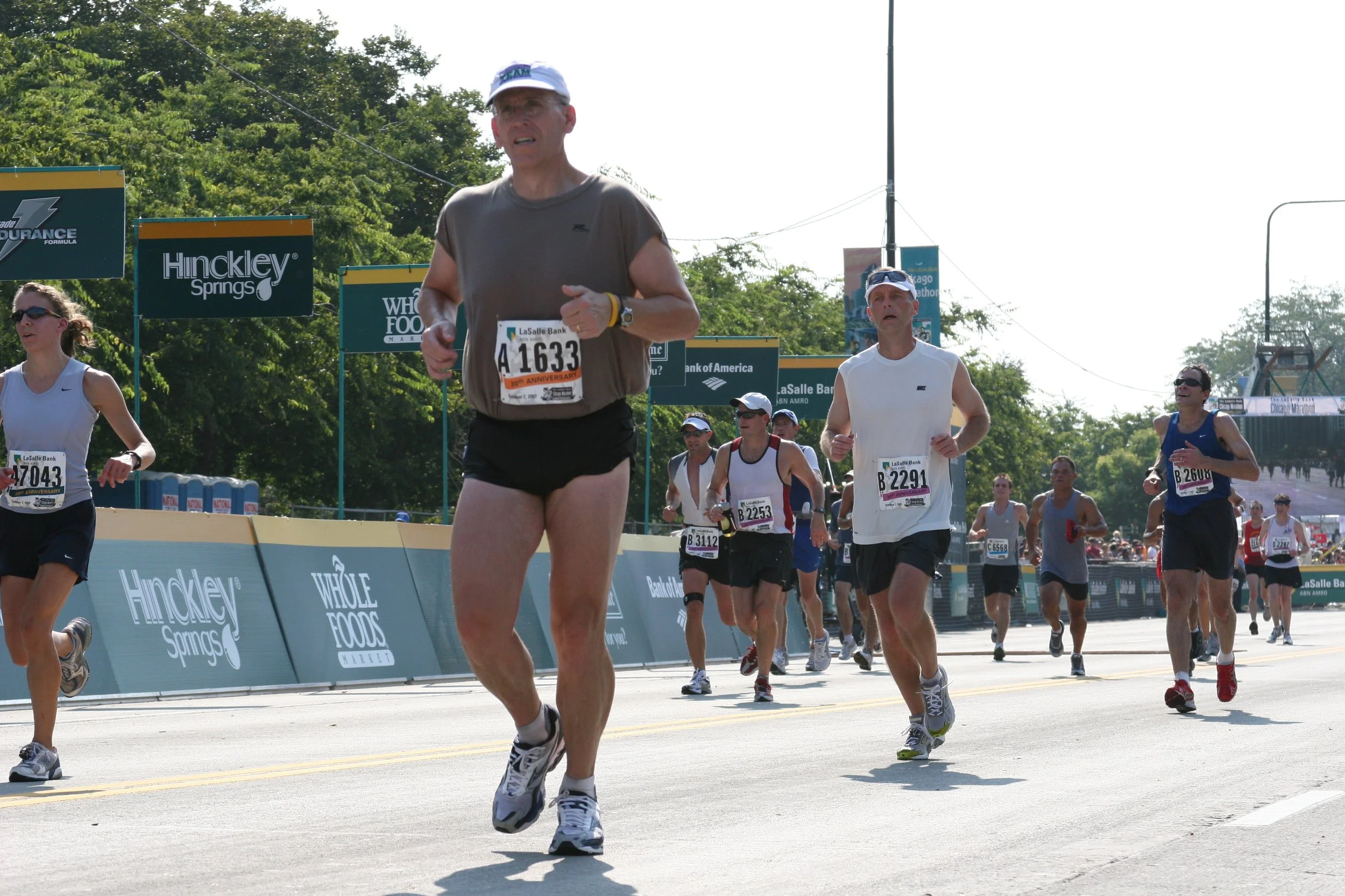 Group of marathon runners participating in the race, running on a street with green trees and banners in the background. The runners are wearing athletic clothing with race bibs.