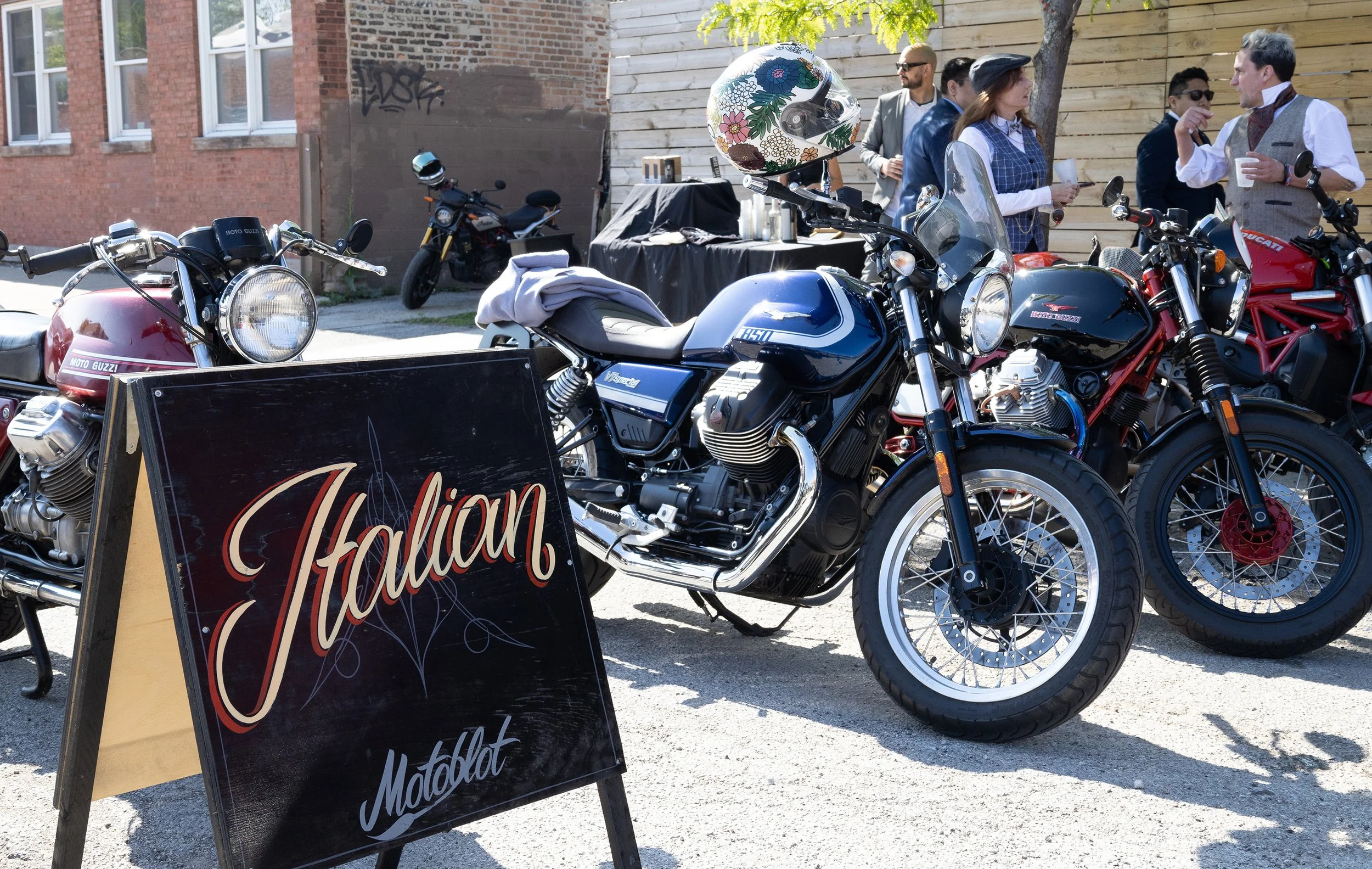 Several motorcycles parked outdoors near a building with a brick wall and wooden fence, with people socializing nearby, and a black sign with red and white lettering that reads 'Italian.'