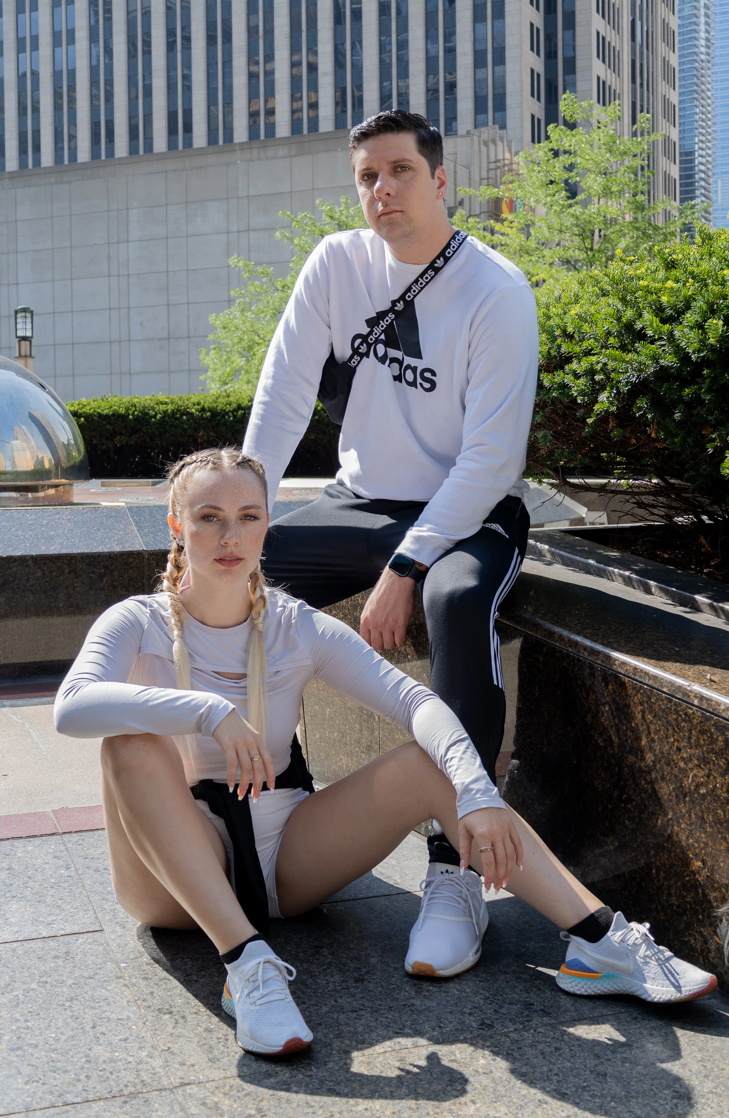 Two young adults, a woman sitting on the ground with her legs crossed and a man sitting on a bench, outdoors in an urban area with tall buildings and green trees in the background. They are dressed in athletic clothing.