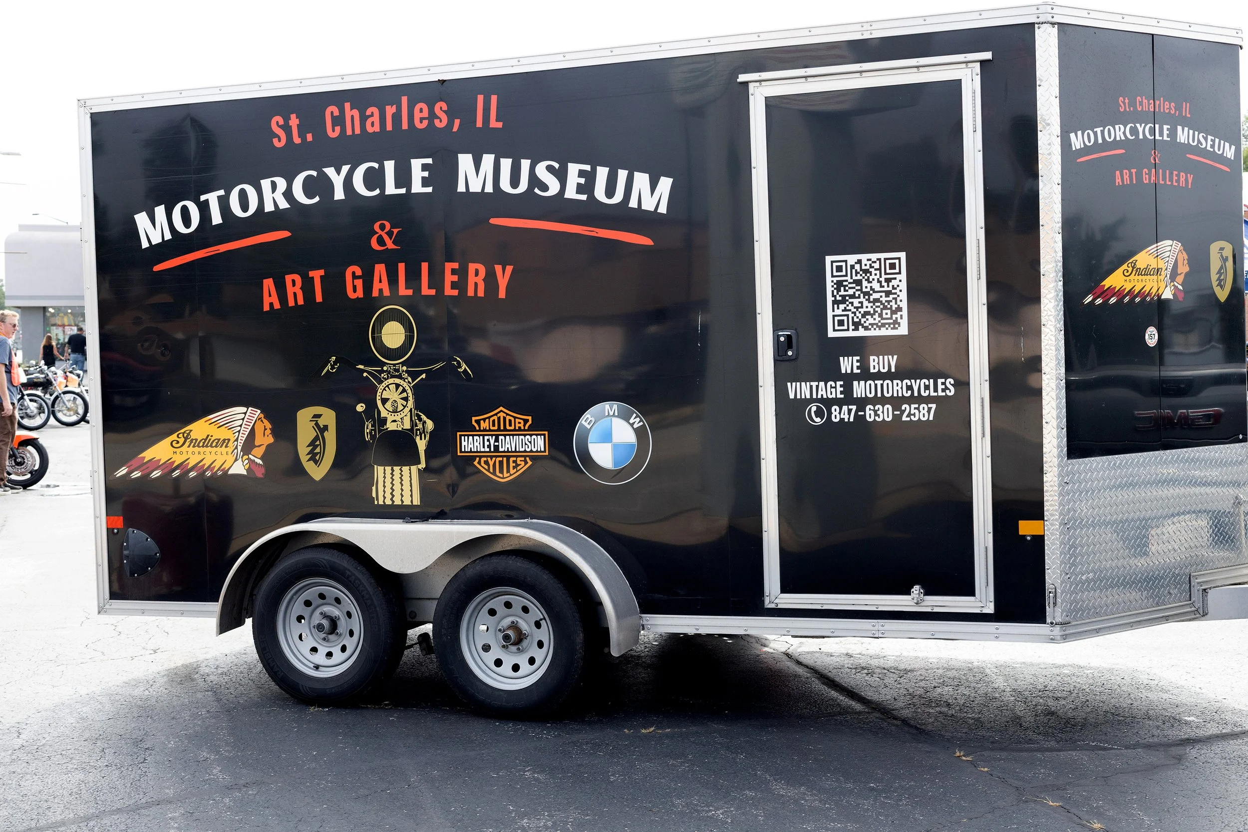 Black trailer truck with signage for St. Charles, IL Motorcycle Museum and Art Gallery, featuring logos of Indian Motorcycle, Harley-Davidson, and BMW, parked on a paved surface.