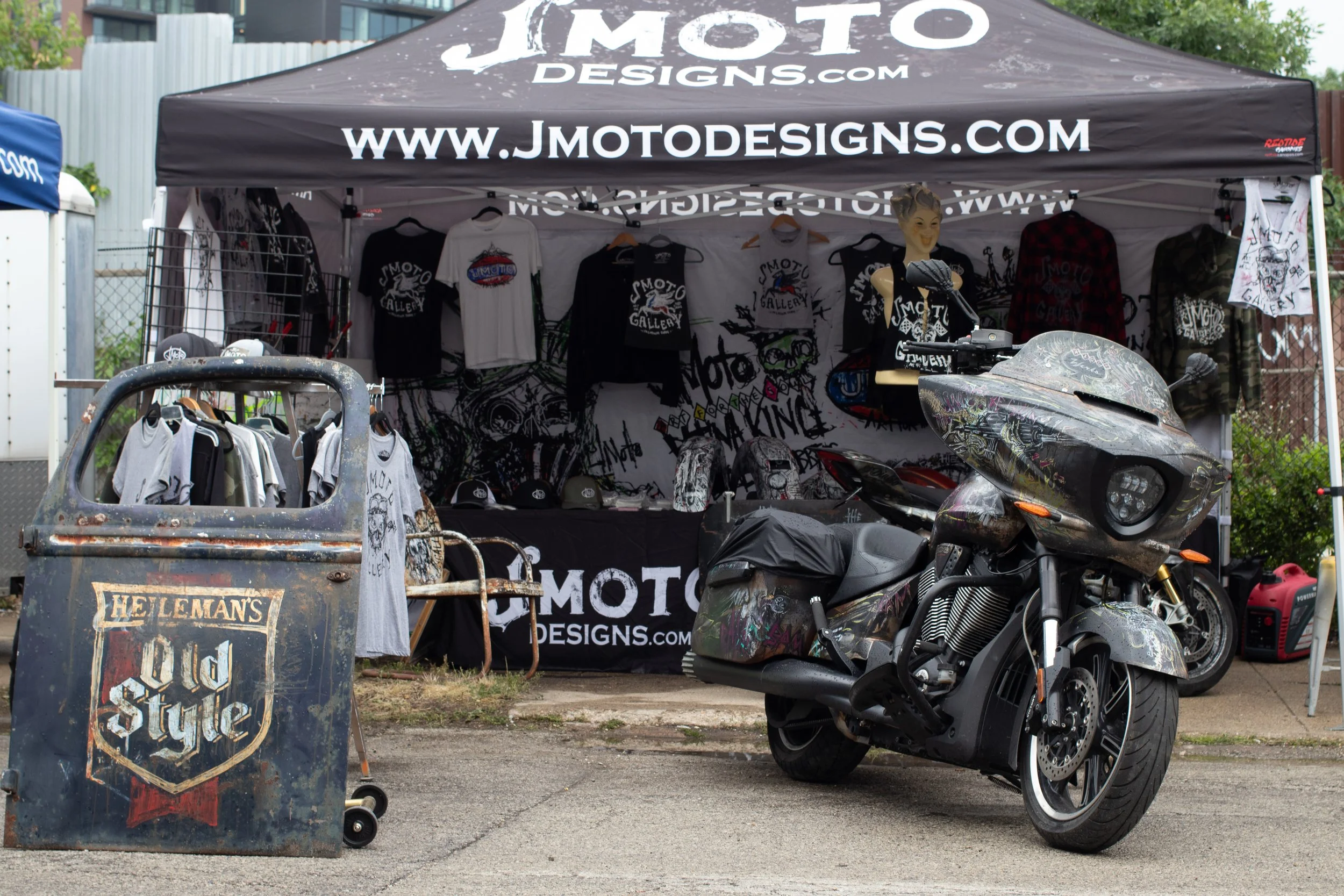 A motorcycle parked next to a black canopy tent with the website 'www.jmotdesigns.com' printed on it. The tent displays motorcycle-themed merchandise such as shirts and hats. In front, there is a rusted metal cart with the words 'Hellman's Old Style'
