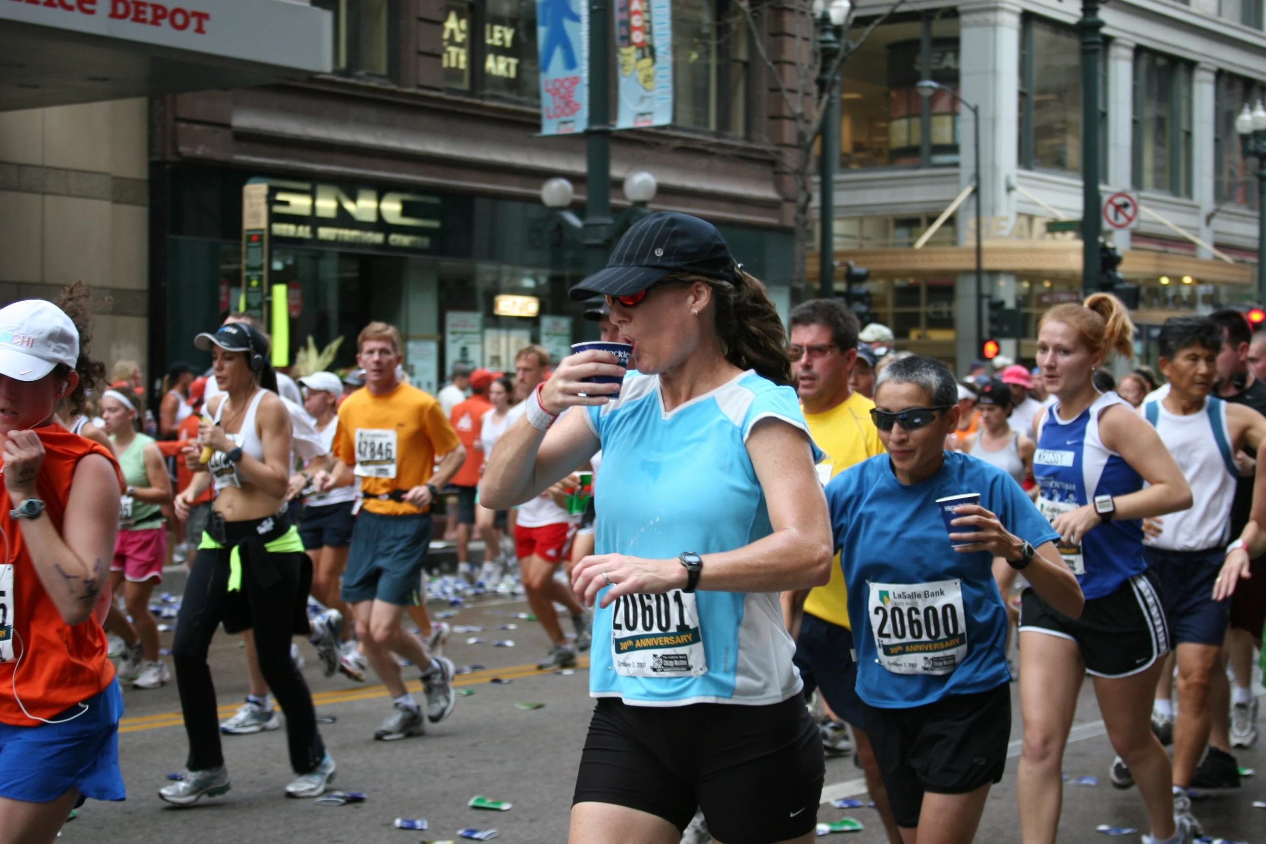 A group of runners participating in a race or marathon on a city street, with some drinking water and others running, surrounded by buildings and spectators.