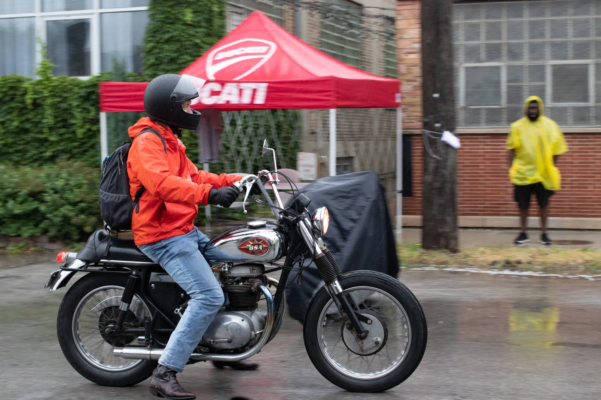 A person riding a black vintage motorcycle on a wet street, wearing a black helmet, orange jacket, and blue jeans. In the background, a red canopy with the word "CALI" and an emblem, a person in a yellow raincoat and dark clothing standing near a bri