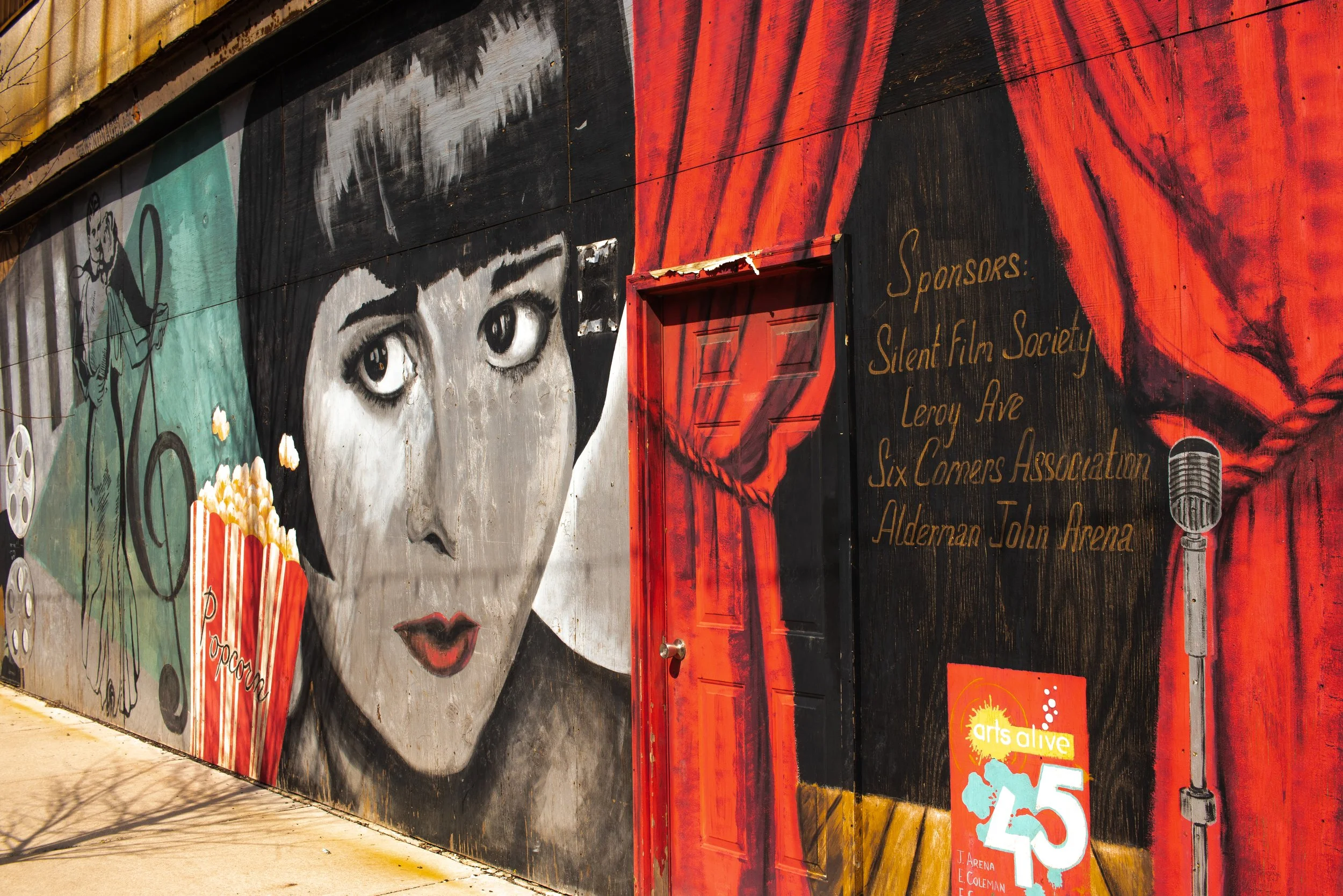 A mural depicting a woman's face in black and white, with large expressive eyes, red lips, and short dark hair. To the right of her face is a red curtain painted on the wall. Next to the curtain, there is a black background with gold text listing spo