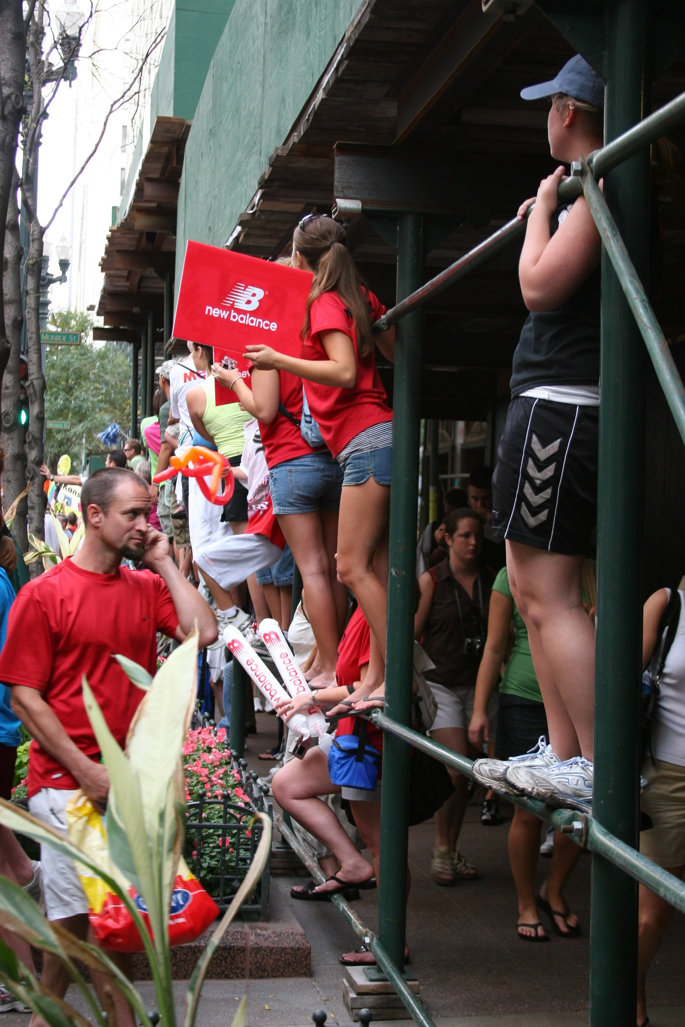 People standing on a scaffolding platform along a city sidewalk during a parade or event. Some are holding signs or balloons. The scene is lively with onlookers and decorations.