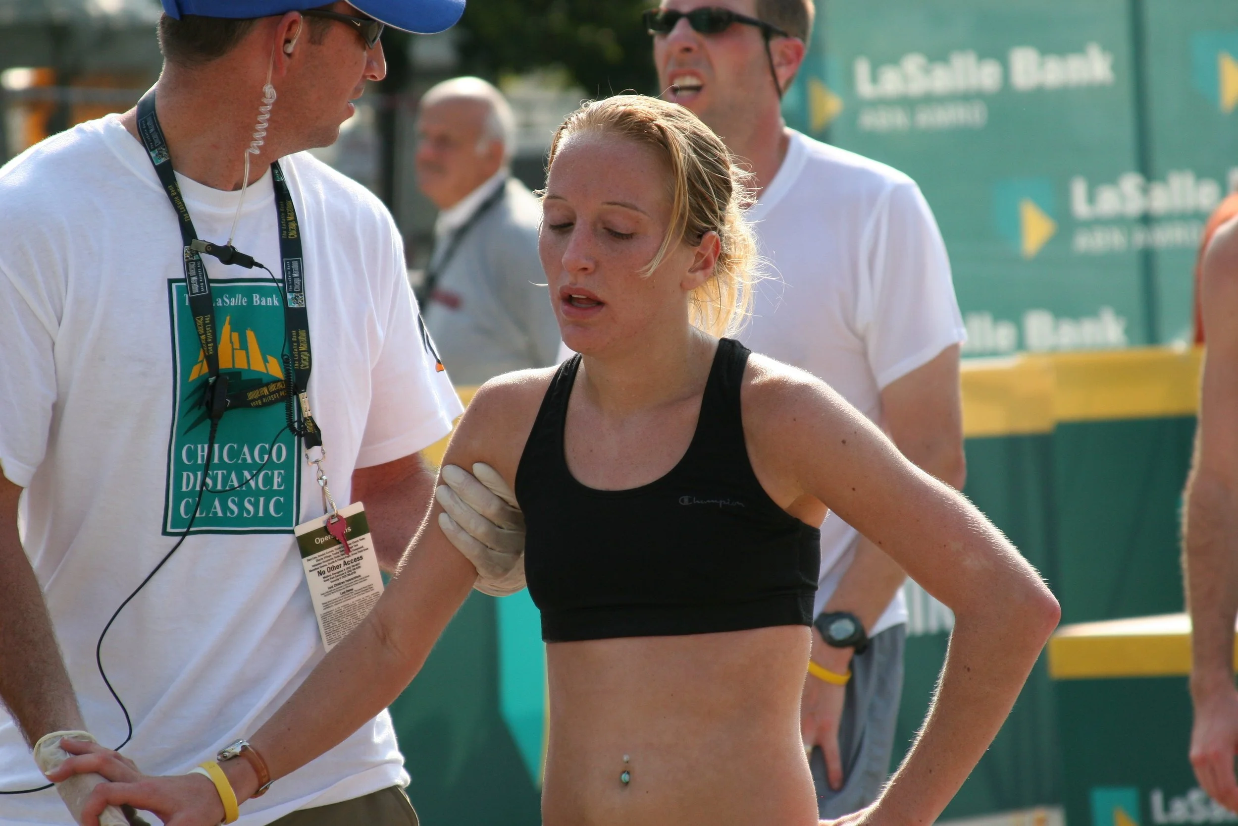A female runner with a stomach piercing and wet hair receives assistance from a man at a marathon event, with other runners and event signage in the background.