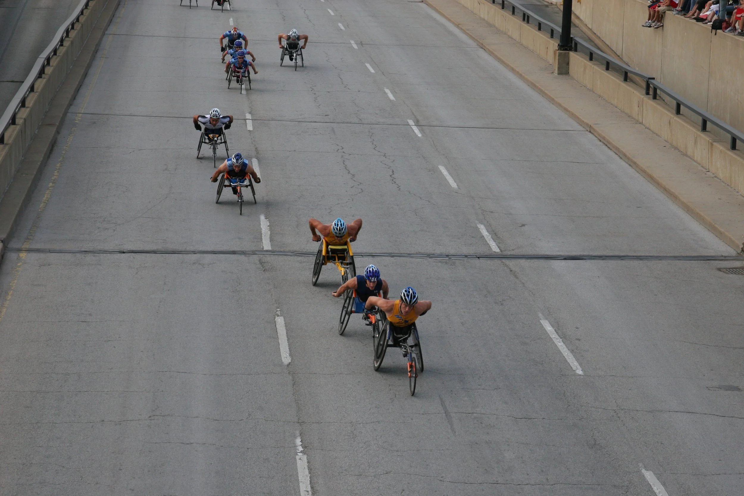 A group of people in handcycles racing on a closed city street, with spectators sitting along the sidewalk.
