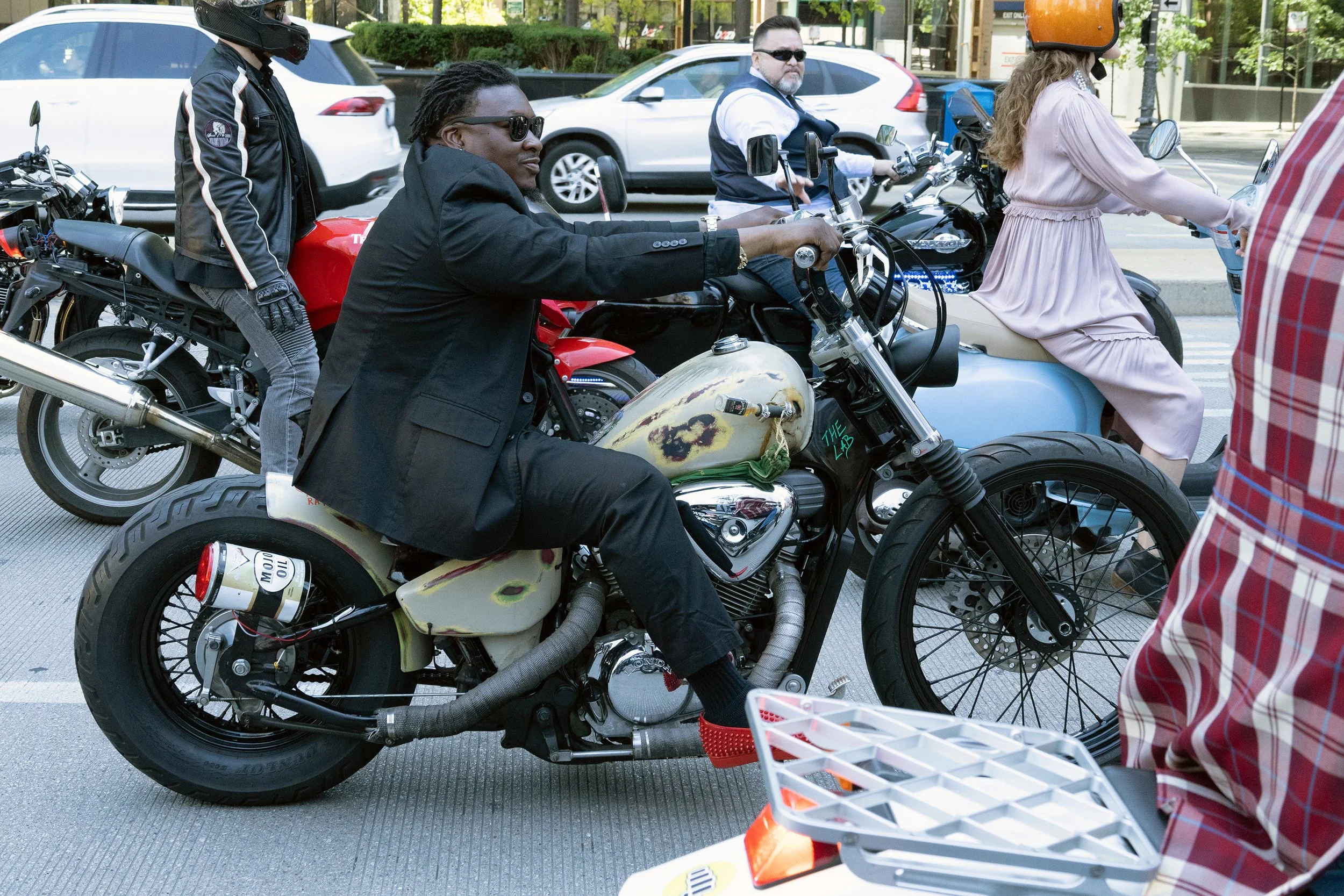 A group of people riding on motorcycles and scooters on a city street. One person is wearing a black jacket, sunglasses, and a helmet, while another is wearing a white shirt and sunglasses. A woman is wearing a light pink dress and a helmet. There ar