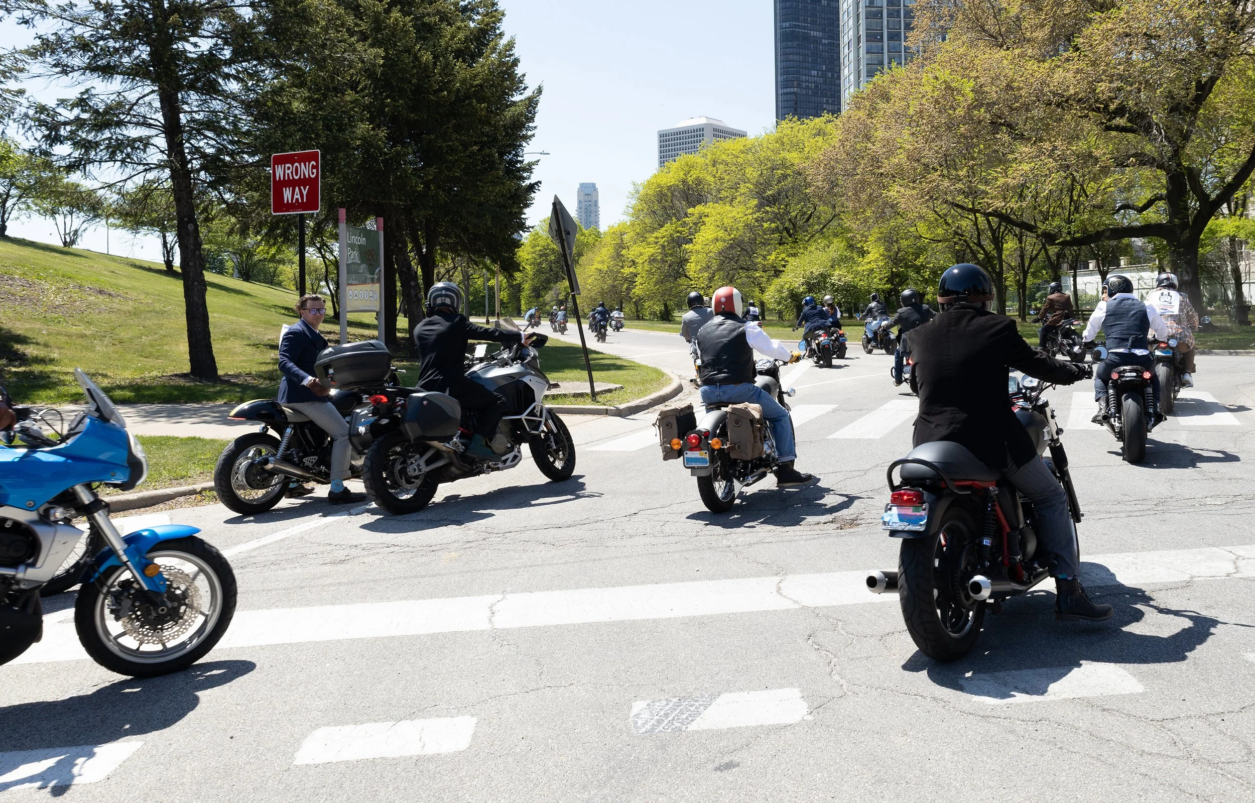 Group of motorcyclists riding on a city street with trees and tall buildings in the background.