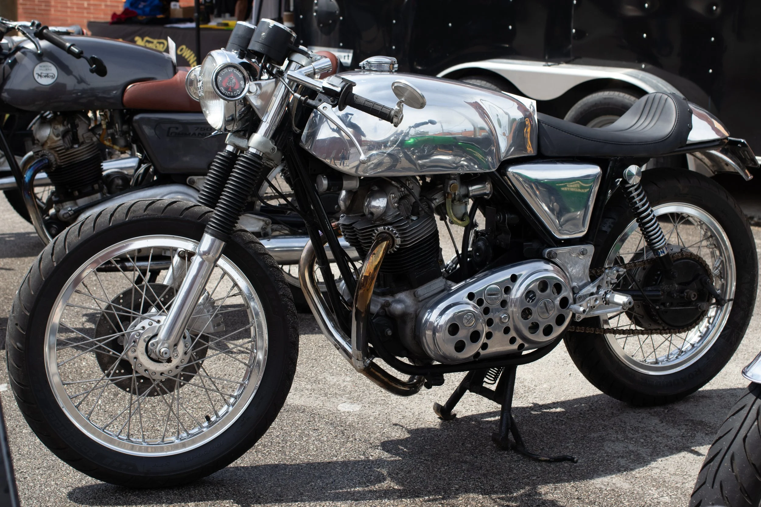 Close-up of a vintage motorcycle with chrome and black parts, parked outdoors. In the background, there is a black vehicle and a red brick building.