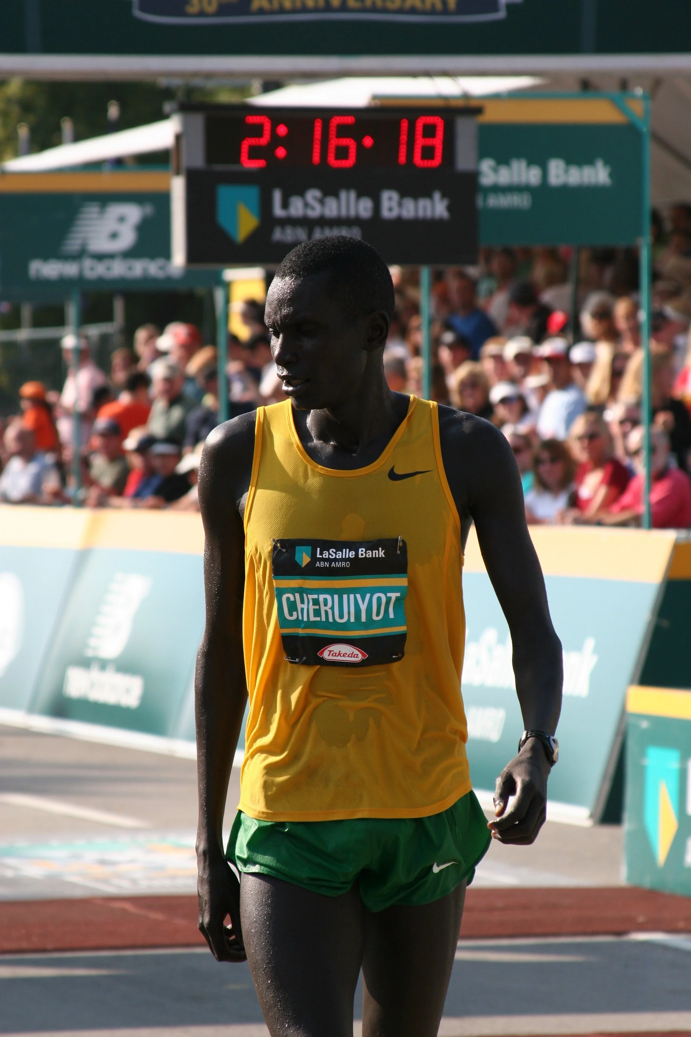 A male marathon runner in a yellow and green uniform with the name Cheruiyot on bib, running during a race at LaSalle Bank with only a few seconds remaining on the digital clock showing 2:16:18, amid a crowd of spectators.
