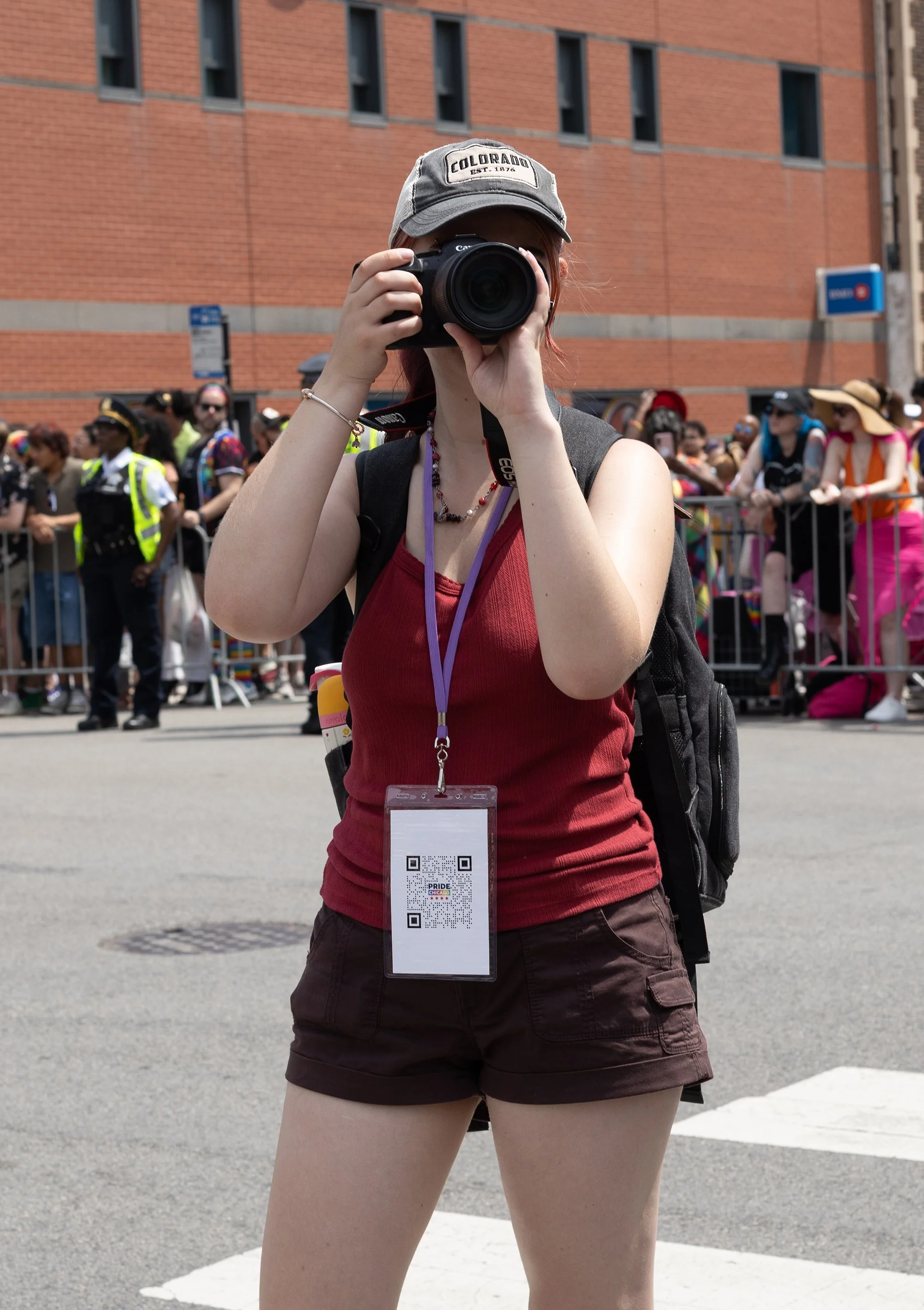 A person in a red top and brown shorts holding a camera, taking a photo at a pride event with a crowd and brick building in the background.