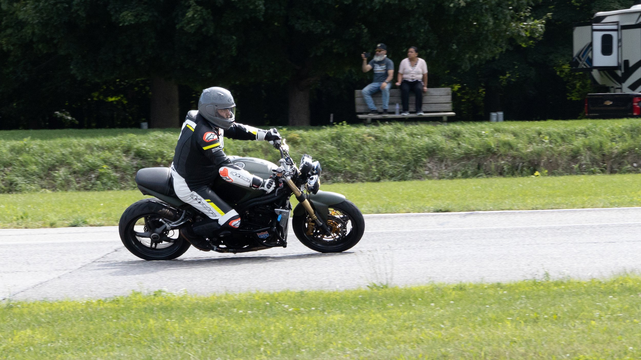 A motorcyclist wearing a black helmet and black riding gear with white and yellow accents is riding a black motorcycle on a road. In the background, two people are sitting on a wooden bench, with a man holding a phone and a woman sitting beside him. 
