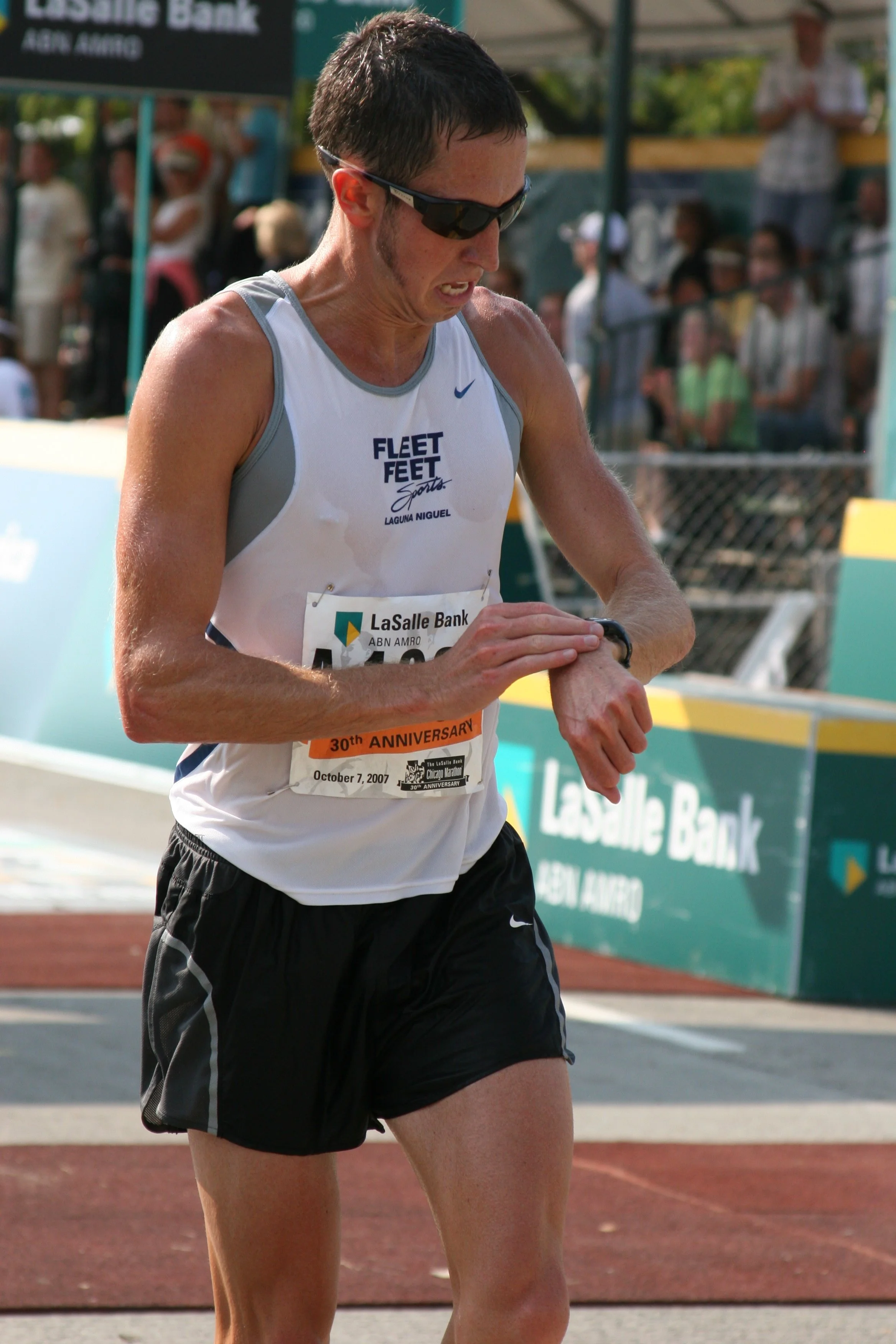 A male runner in a white tank top and black shorts is crossing a finish line. He is wearing sunglasses and a watch, with a tense expression, and a crowd and banners are visible in the background.