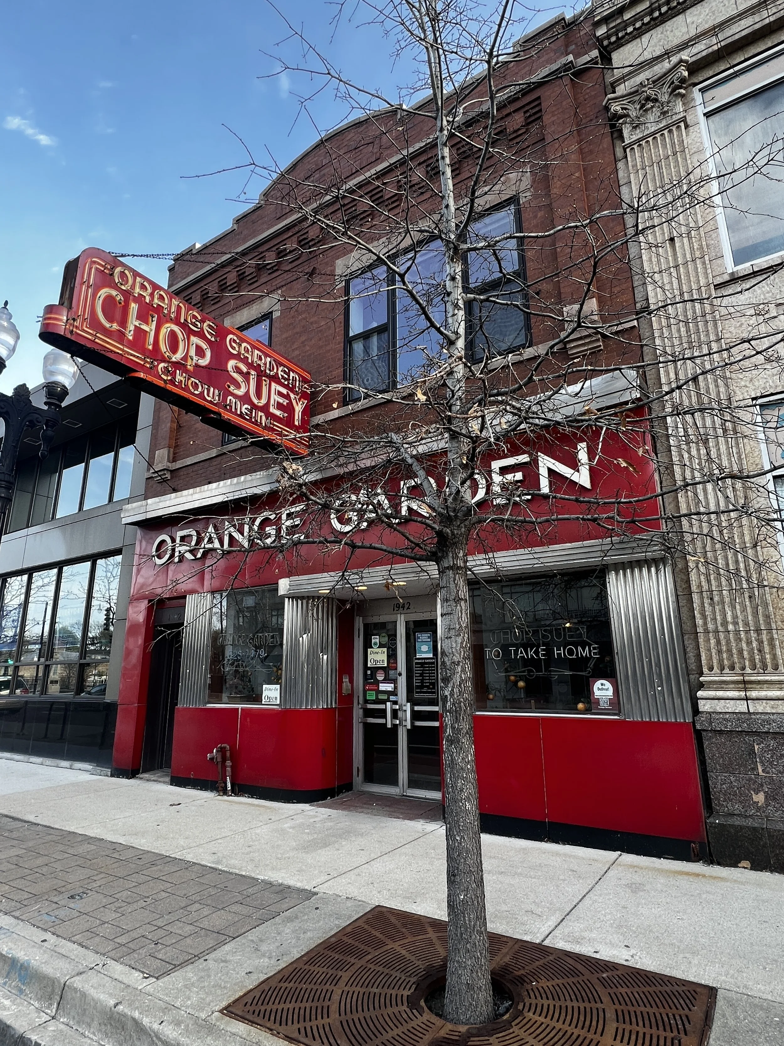 A storefront with red and gray exterior walls, a neon sign reading 'Orange Garden Chop Suey & Chow Mein,' and a sign below indicating 'Takeout & Delivery.' There is a leafless tree in front of the store and a sidewalk with a street drain.