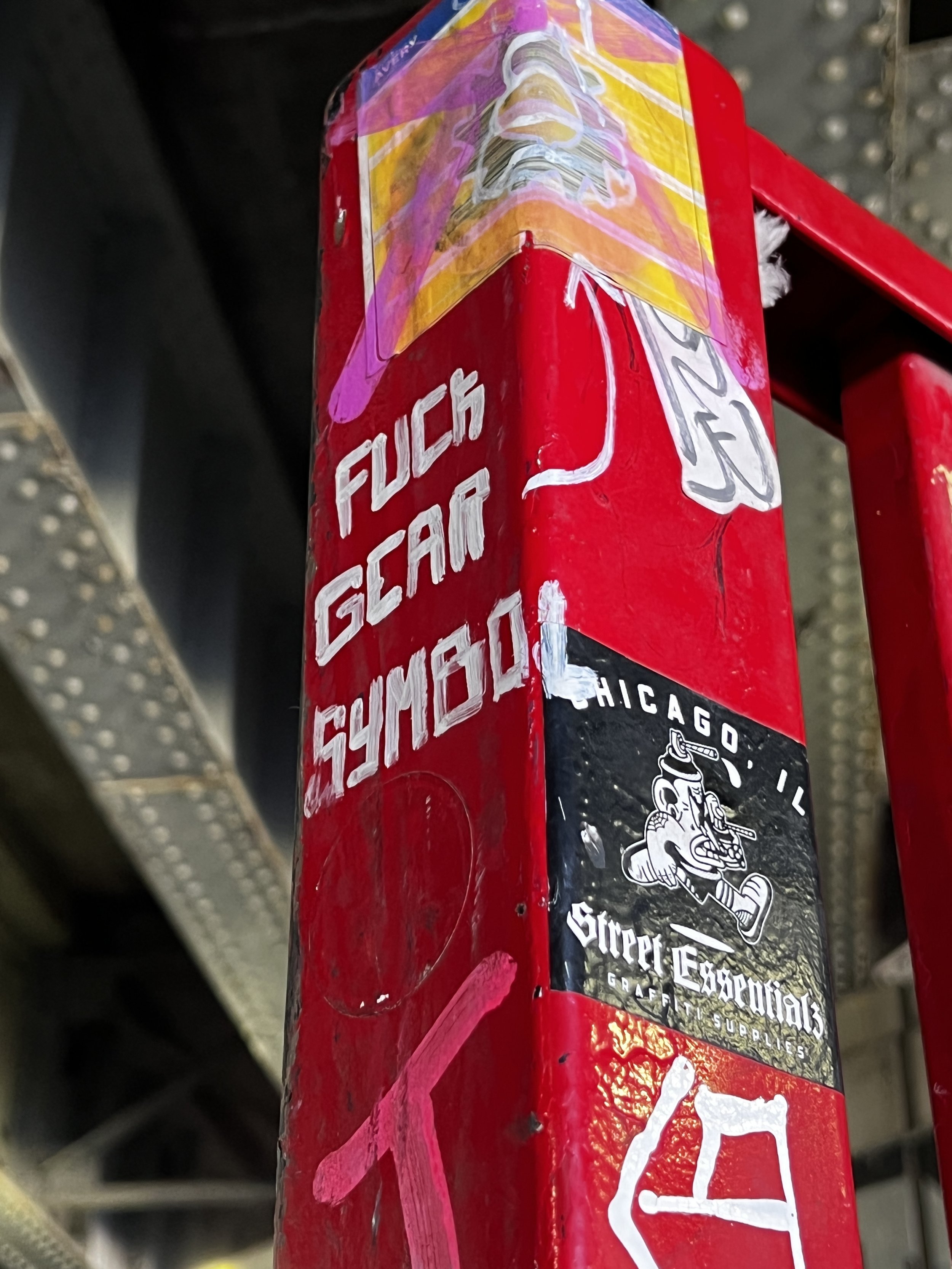 Close-up photo of a red bicycle frame with a colorful sticker depicting a stylized face with long hair. The frame has white graffiti-style text and logos, including a stylized skull with a spear. The background shows a metal surface with small rivets