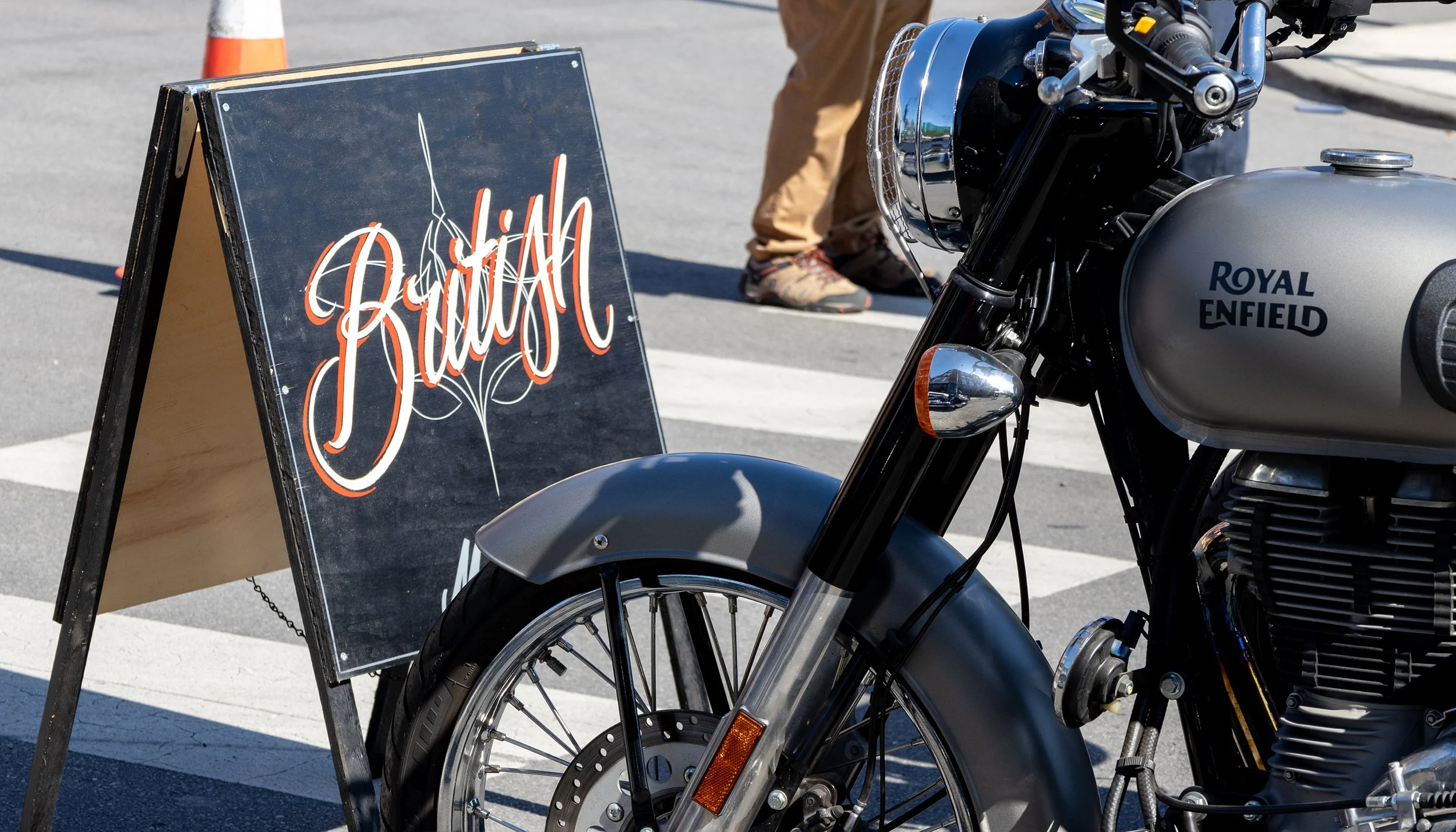 A close-up of a Royal Enfield motorcycle parked on the street with a black chalkboard sign that has a decorative white and orange script reading "Bike" on it. A person's legs and shoes are visible in the background.