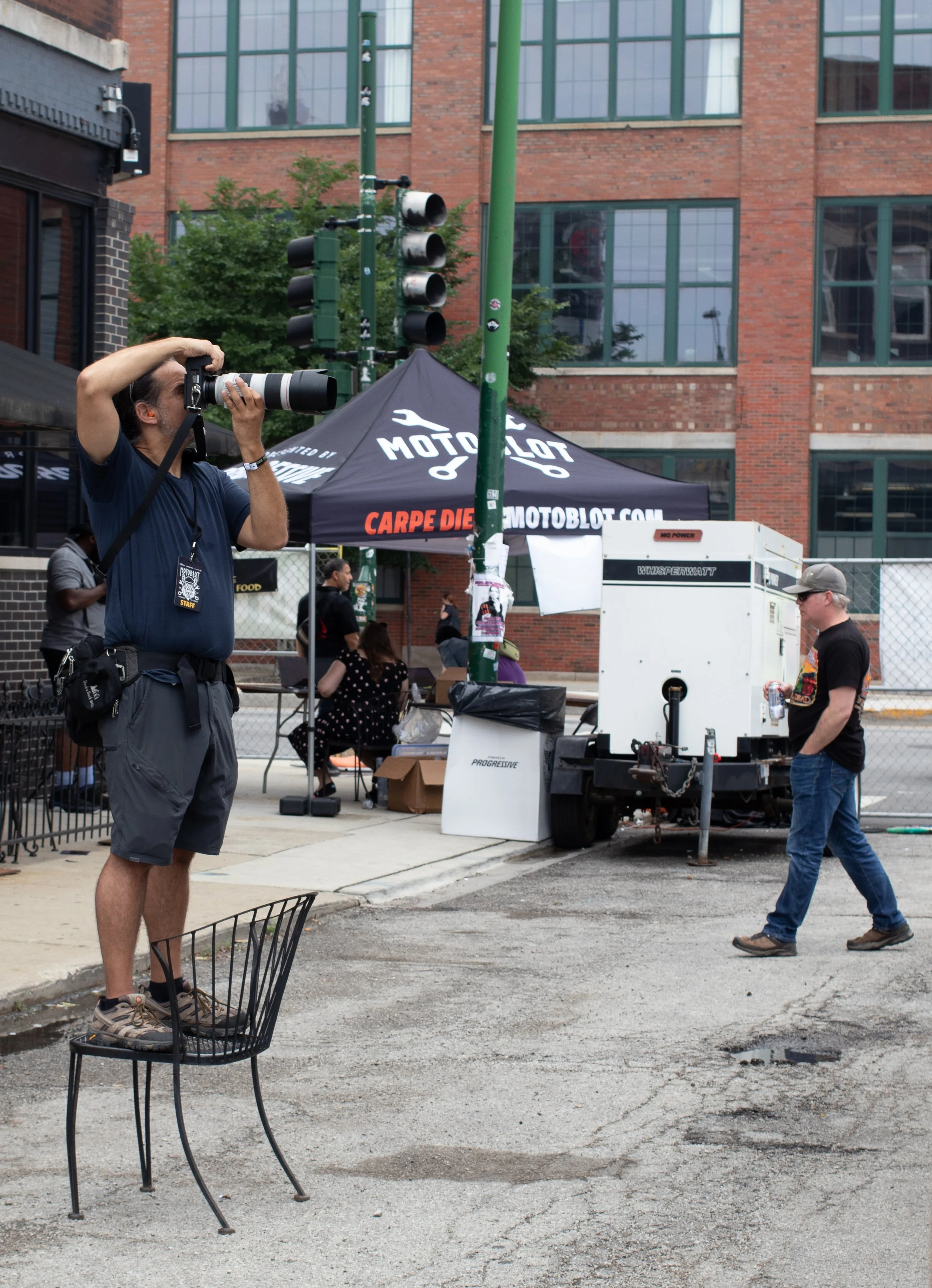 A man with a camera taking a photo at an outdoor event in front of a tent with the MotoBot logo, along with several people and equipment, in an urban setting with brick buildings and green lampposts.