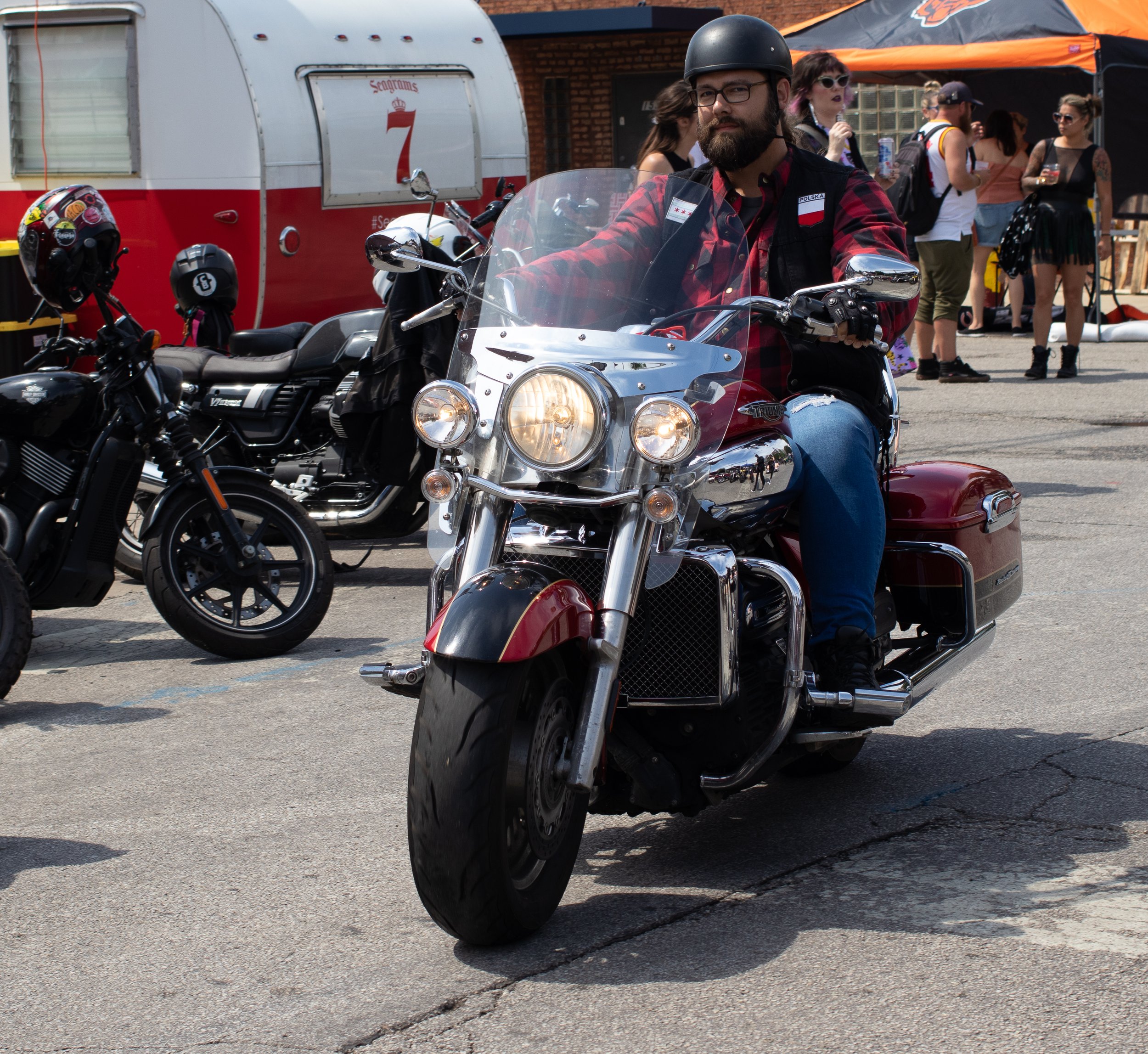 A man with a beard and glasses riding a red motorcycle on a street, wearing a black helmet and a red flannel shirt. There are people in the background, some holding drinks and wearing sunglasses, at an outdoor event.
