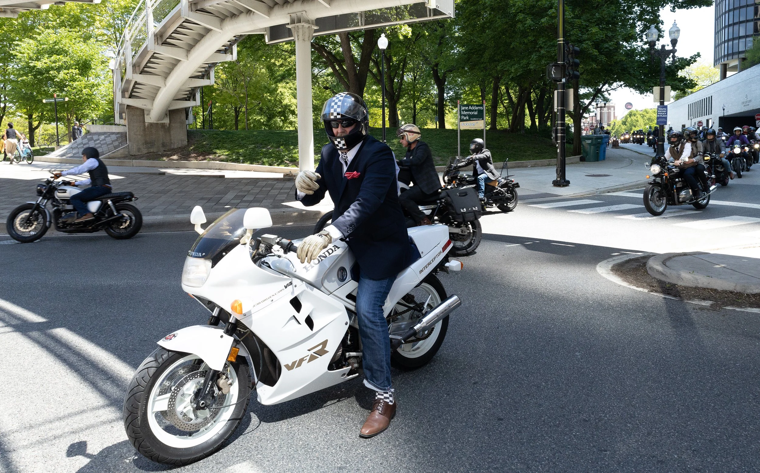 Man in business suit and helmet riding a white Honda motorcycle at an intersection with other motorcyclists and pedestrians nearby. Green trees and urban buildings in the background.
