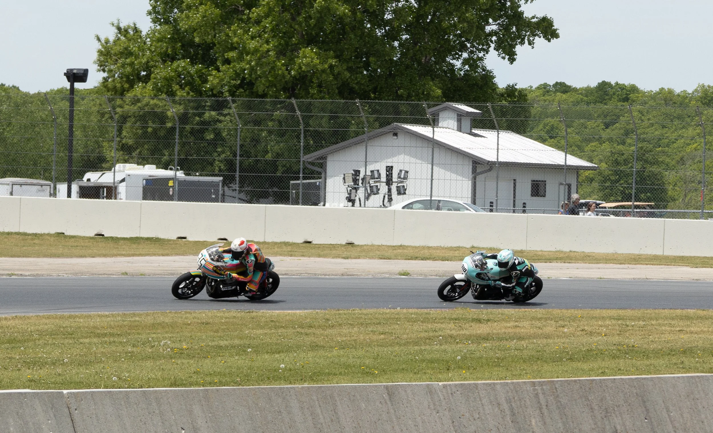 Two motorcycle racers in colorful racing suits and helmets speed around a race track, with a fence, a small building, and some equipment in the background.