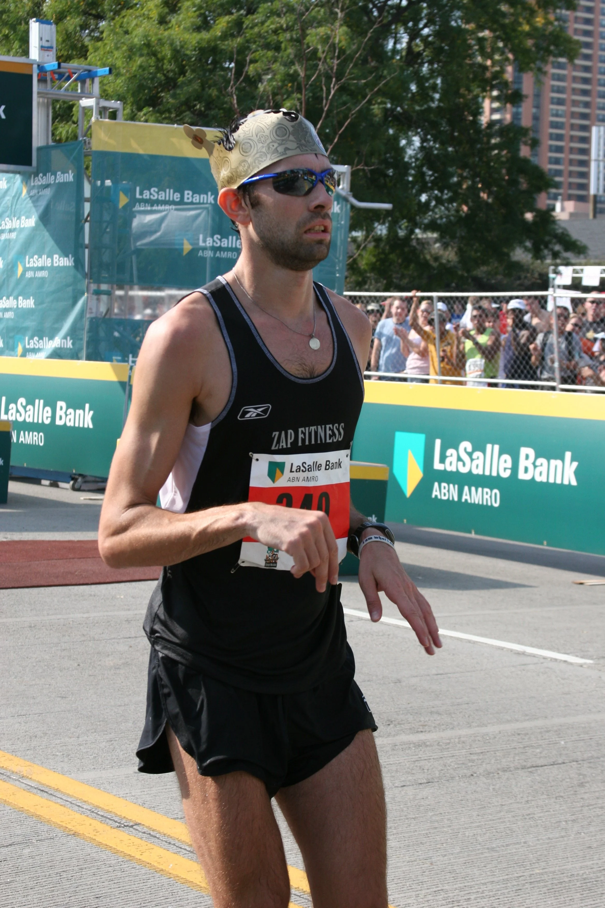A male marathon runner in black athletic attire, wearing sunglasses and a beige head covering, running on a city street during a race, with a crowd of spectators behind a fence and lush green trees in the background.