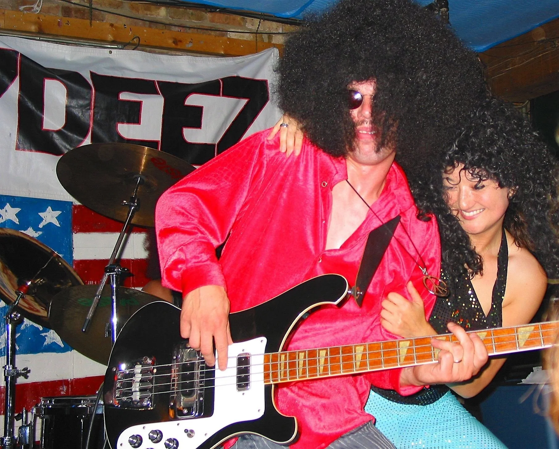 A man and woman dressed in rock and roll costumes, with big afro wigs, playing guitars and enjoying music on stage with American flag-themed decorations behind them.