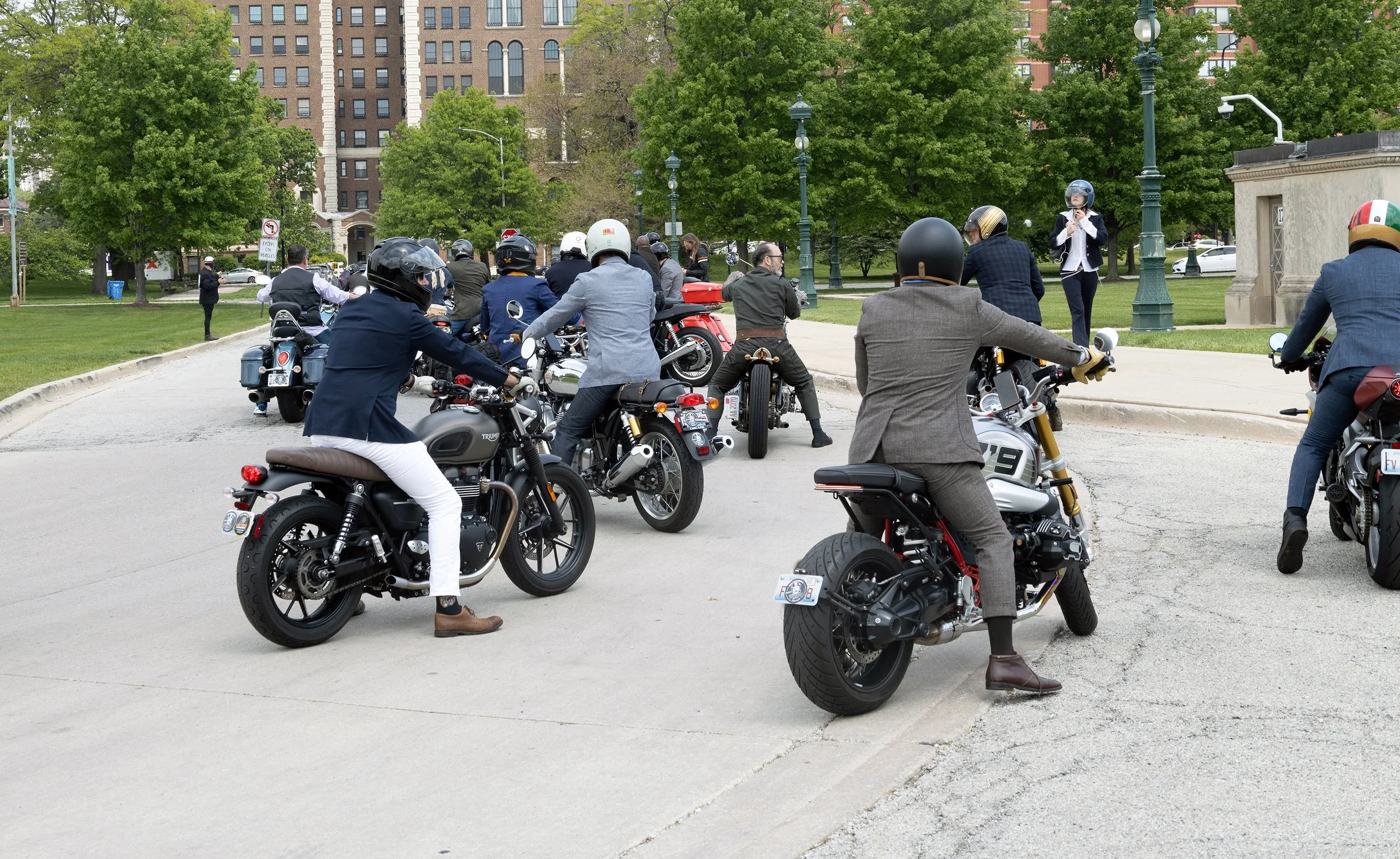 A group of people in business attire, wearing helmets, riding motorcycles and scooters in a city park area with trees and tall buildings in the background. Several individuals are stopped and are looking at each other or towards a speaker.