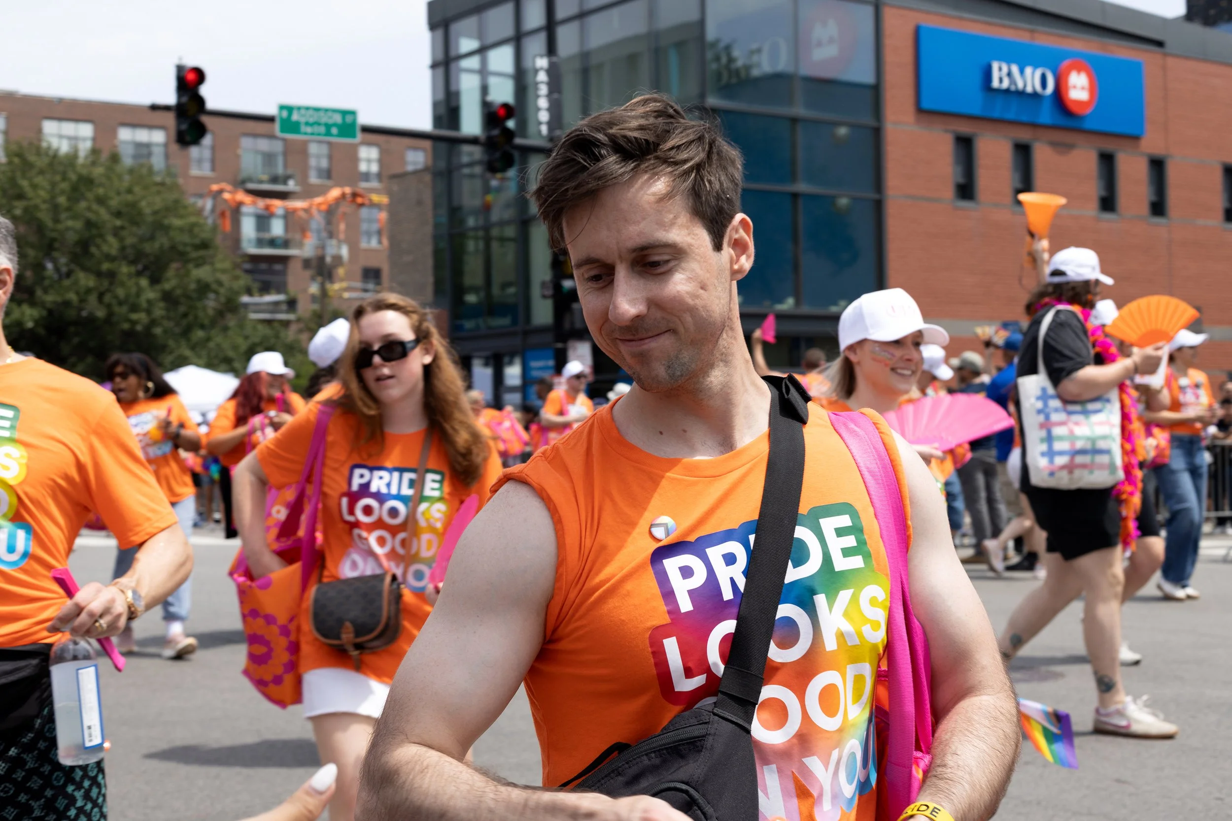 A man with short brown hair wearing an orange Pride T-shirt and a pink backpack participating in a Pride parade, surrounded by other people also in orange attire, in front of a modern building with a BMO logo, traffic lights, and festive decorations.