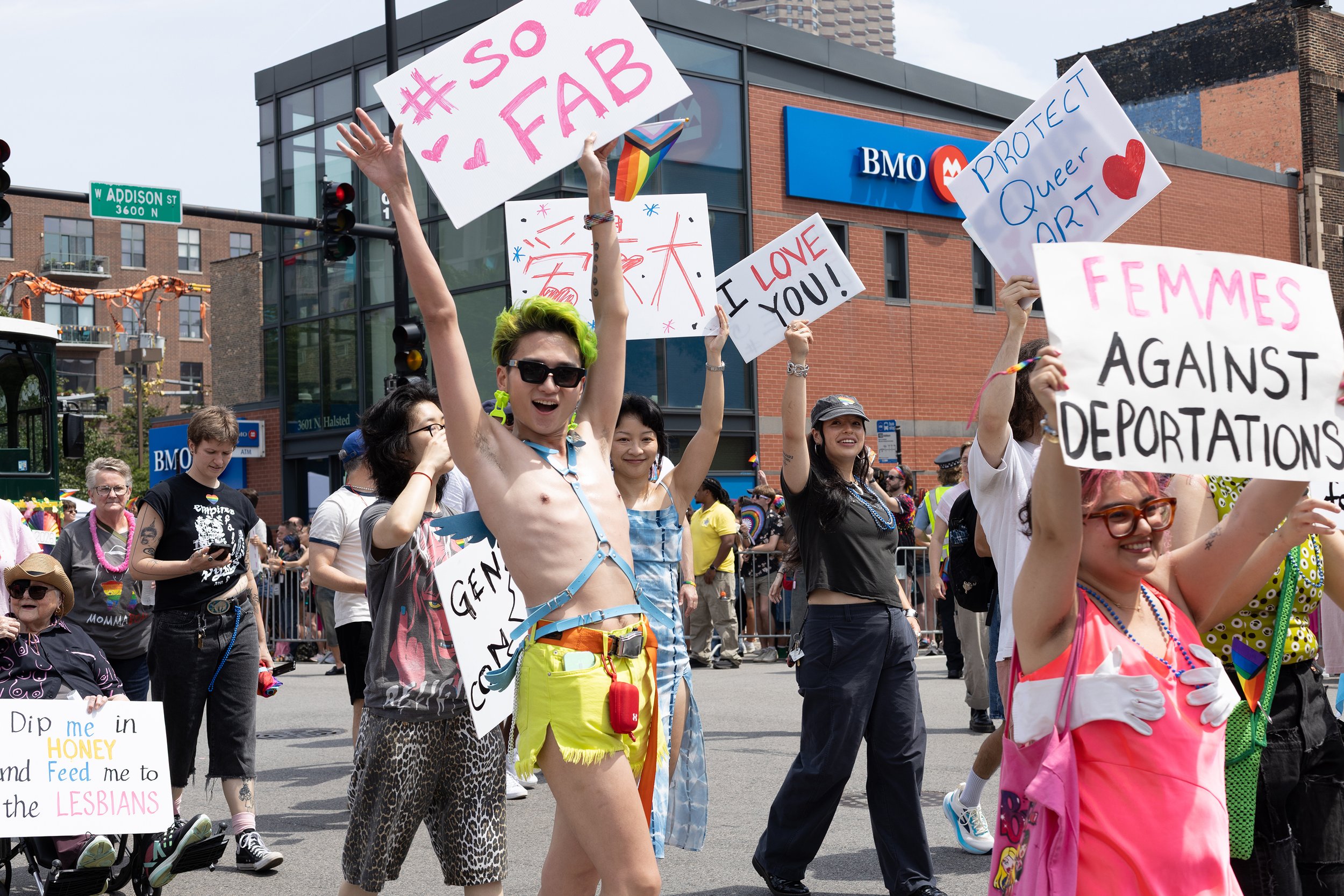 A group of diverse people participating in a pride parade, holding signs supporting LGBTQ+ rights and love, with a backdrop of city buildings and a BMO bank.
