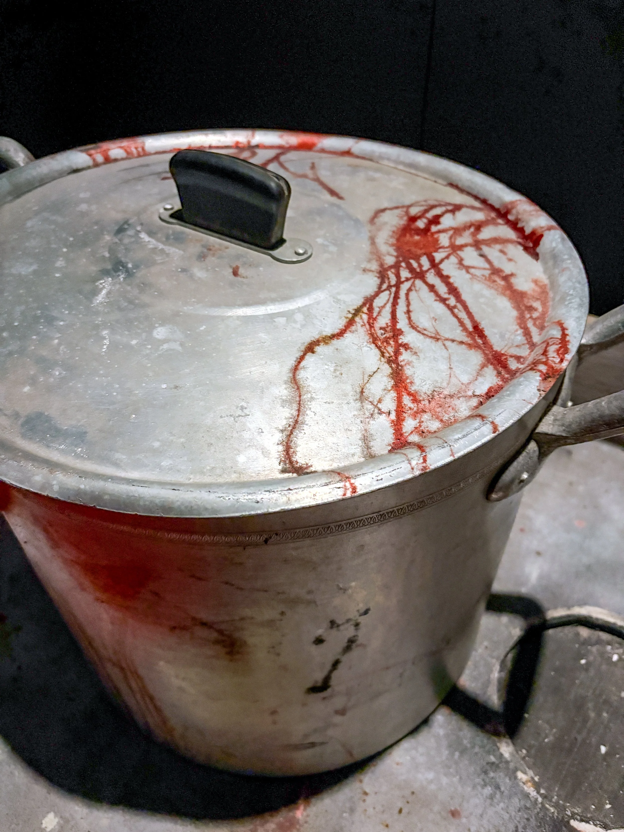 A large, old stainless steel cooking pot with a black handle on the lid, stained with red residue and black smudges, placed on a concrete or stone floor.
