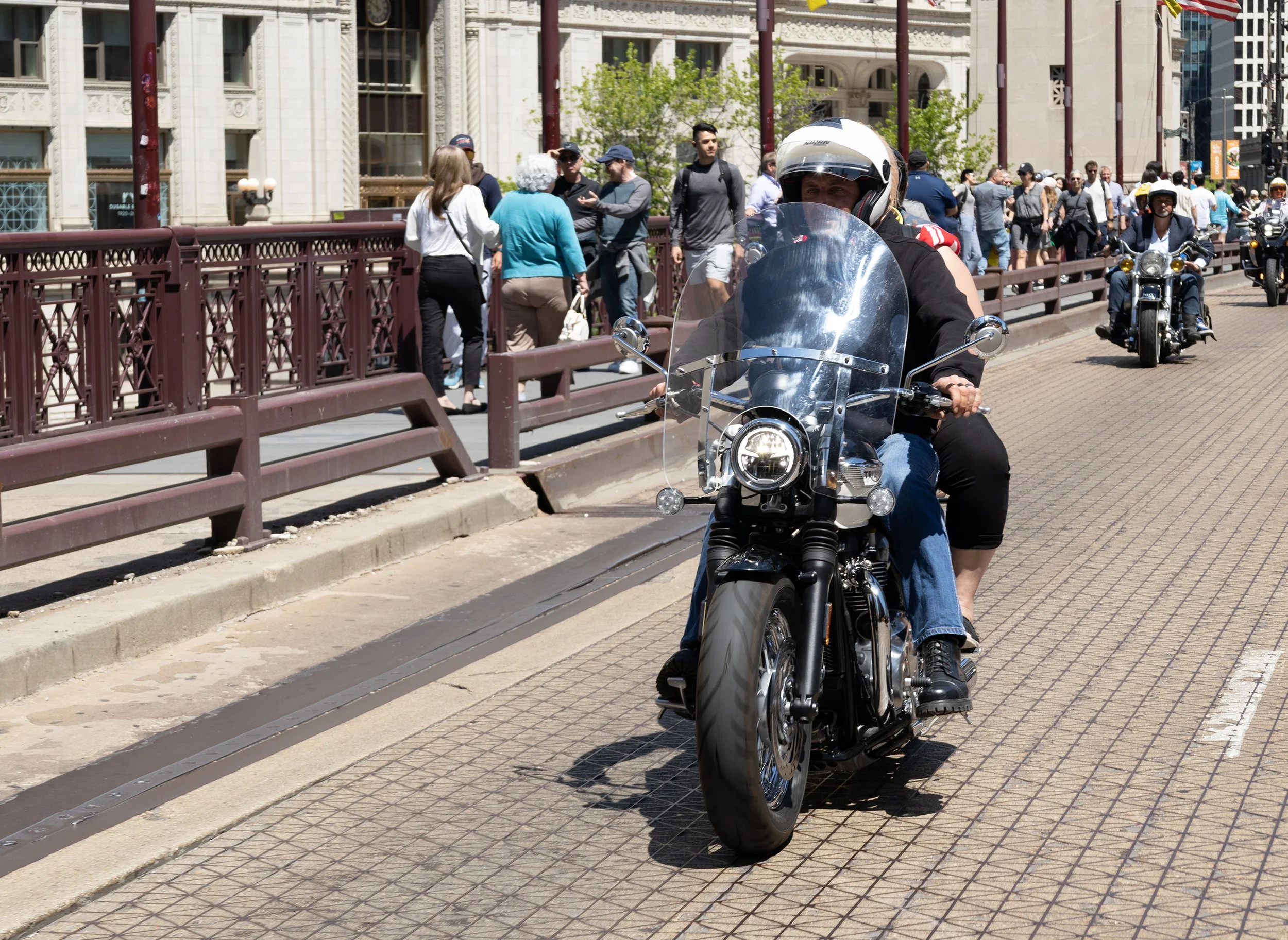 A person riding a motorcycle with a passenger standing on the street. There are buildings and a crowd of people in the background on the city bridge on a sunny day.