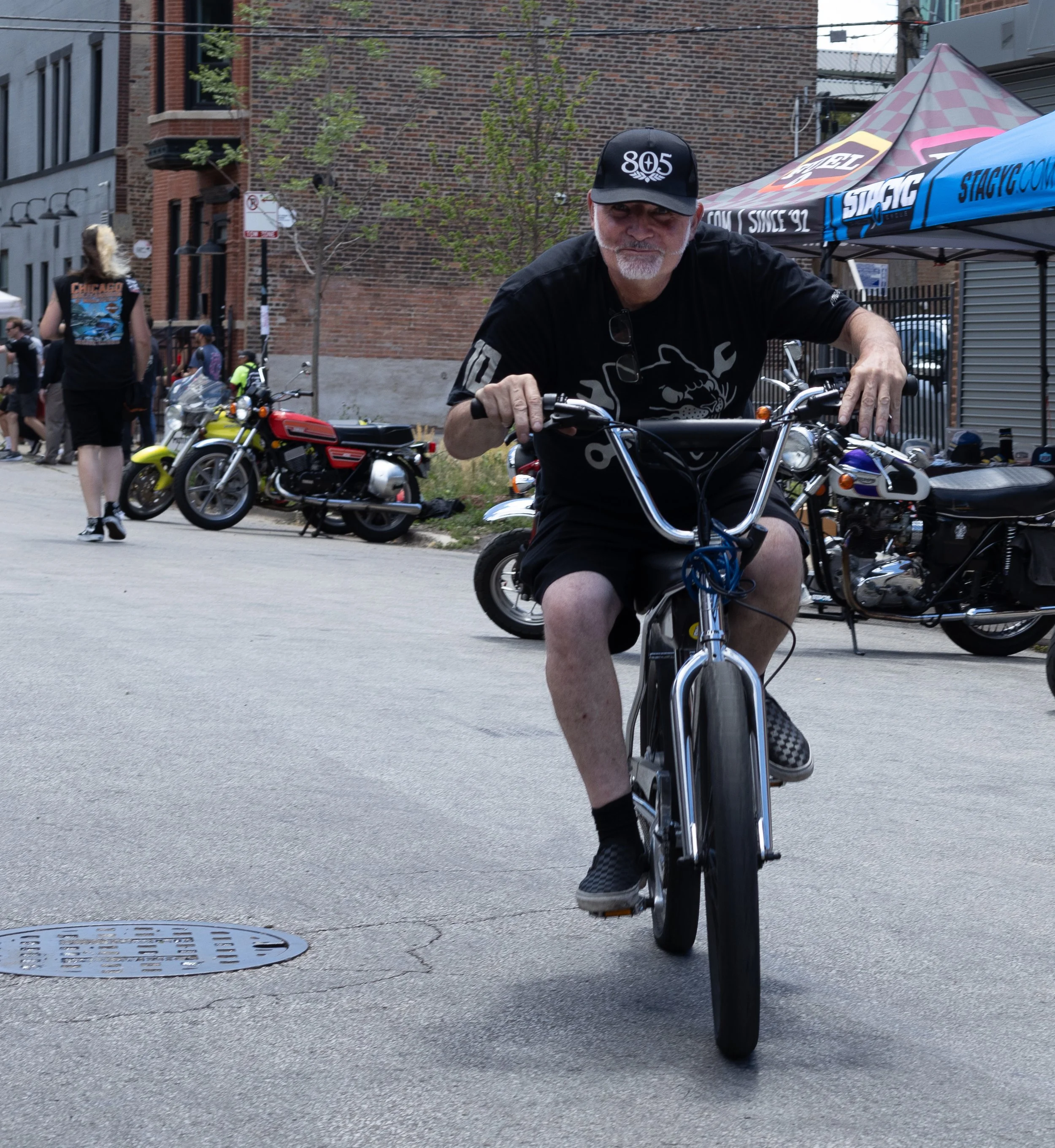 A man riding a bicycle on an urban street with old brick buildings and parked motorcycles in the background. The man has a gray beard, is wearing a black hat with '805' on it, sunglasses on his shirt collar, and a black t-shirt with a graphic design.