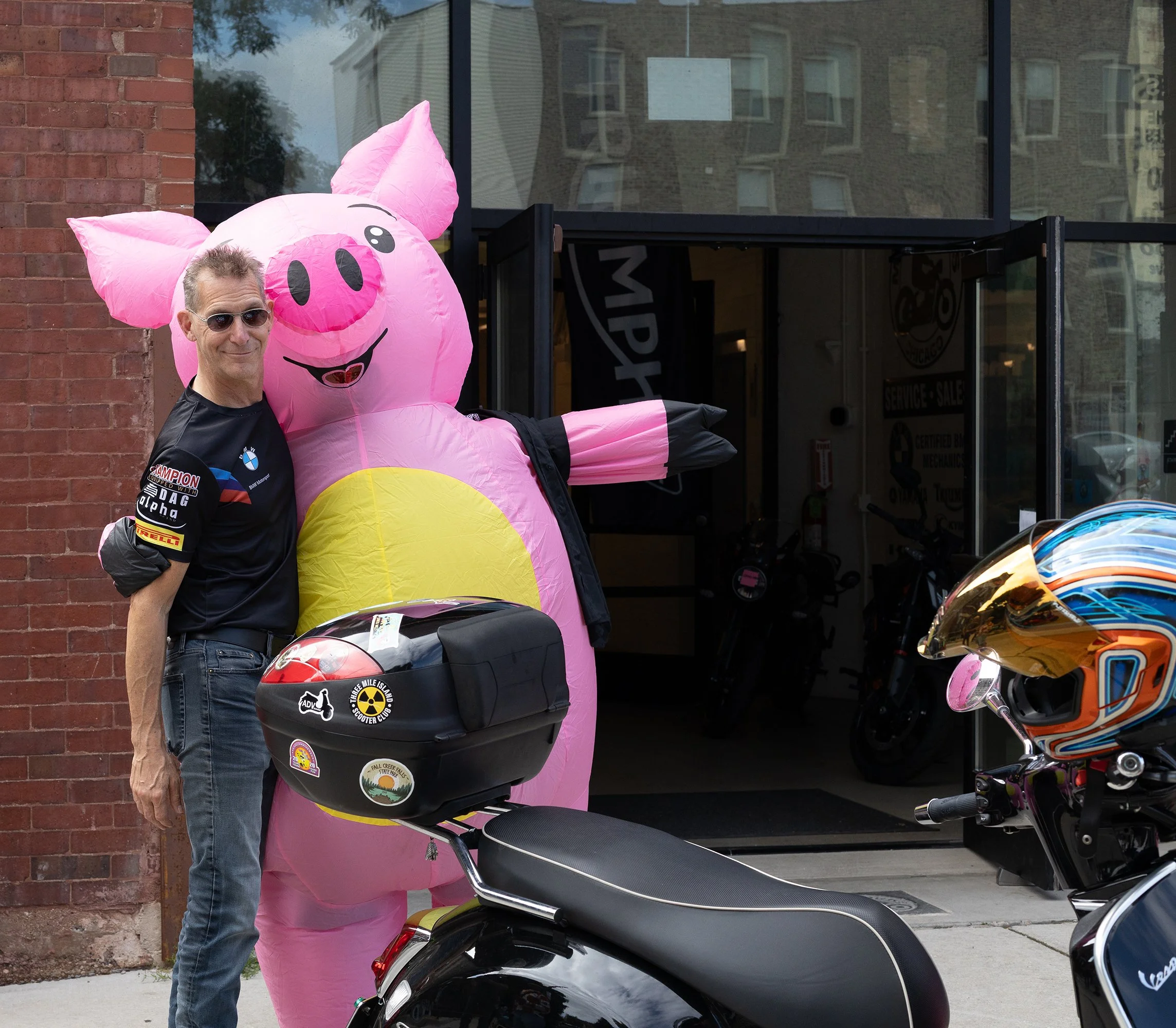 A man wearing sunglasses and a black shirt standing next to a large pink inflatable pig outside a building with glass windows and a brick wall.