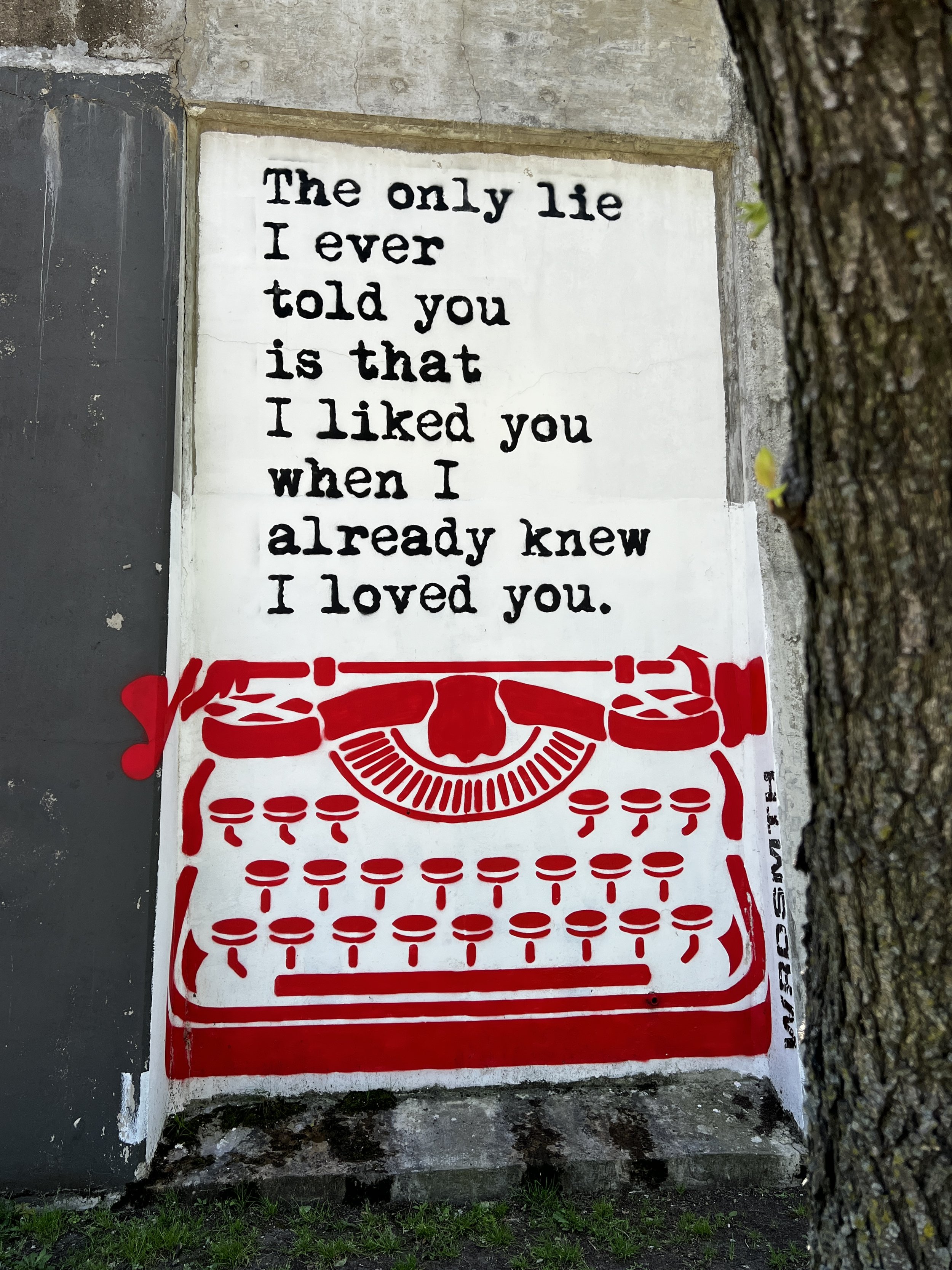 A wall with a white sign featuring black text that reads, "The only lie I ever told you is that I liked you when I already knew I loved you." Below the sign, part of a red design depicting a table with chairs is visible, and a tree trunk is partially