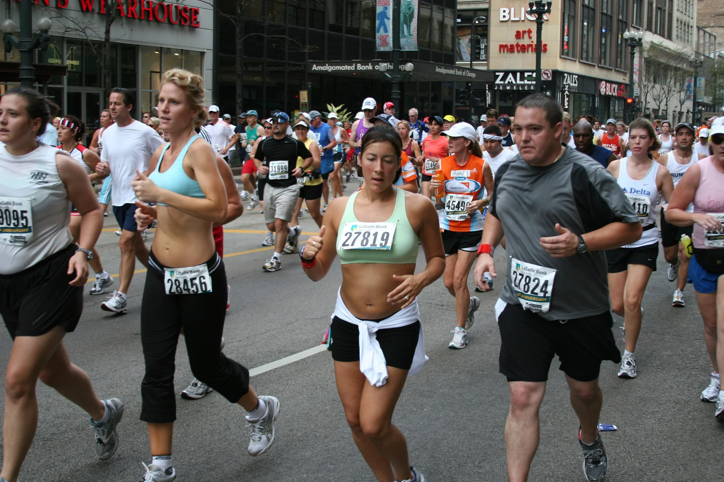 A large group of runners participating in a marathon or race on a city street, with some wearing race bibs, in front of buildings and storefronts.