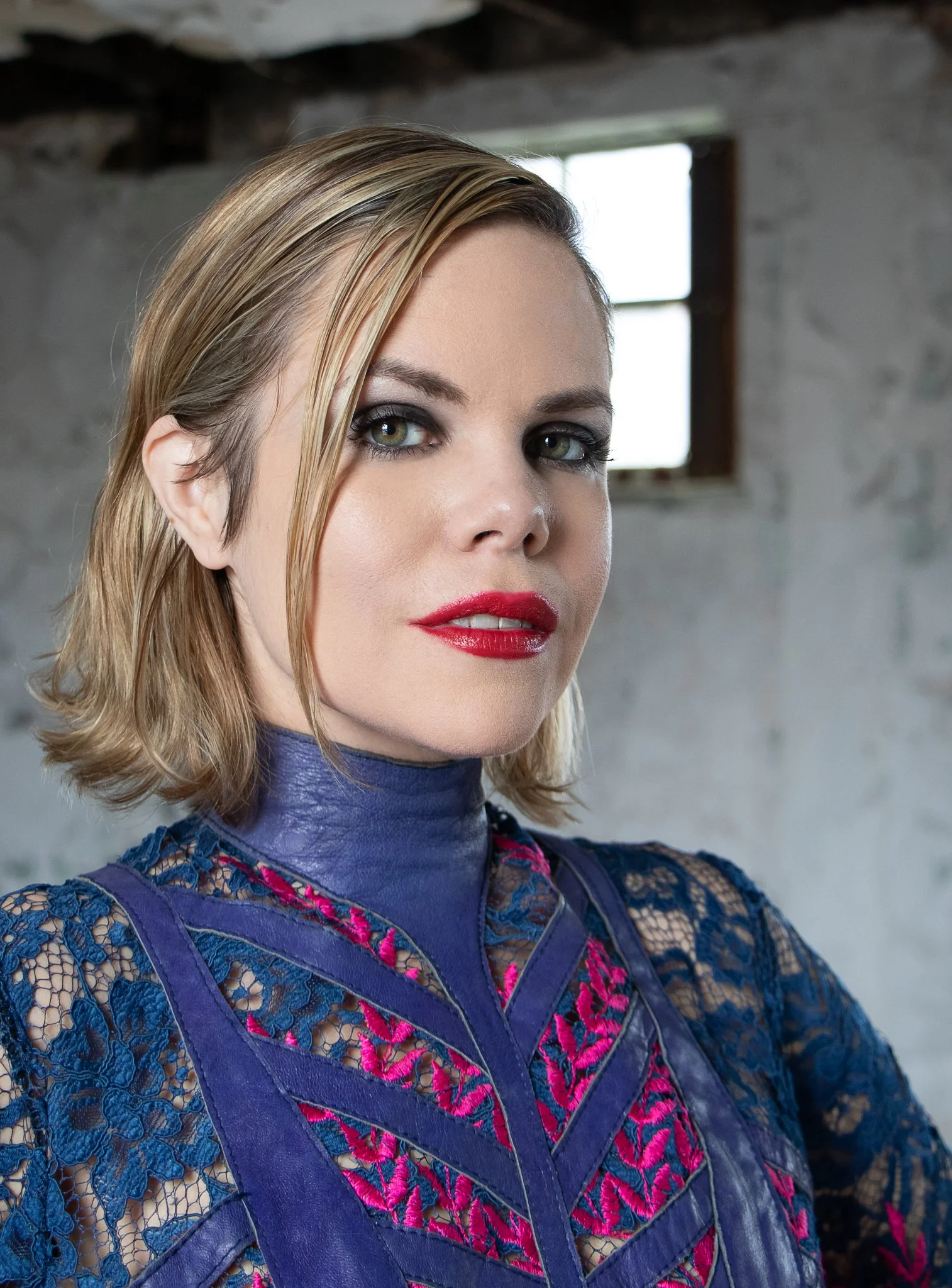 A woman with blonde, shoulder-length hair, wearing a blue lace dress with pink embroidery, standing inside a rustic building with a window in the background.