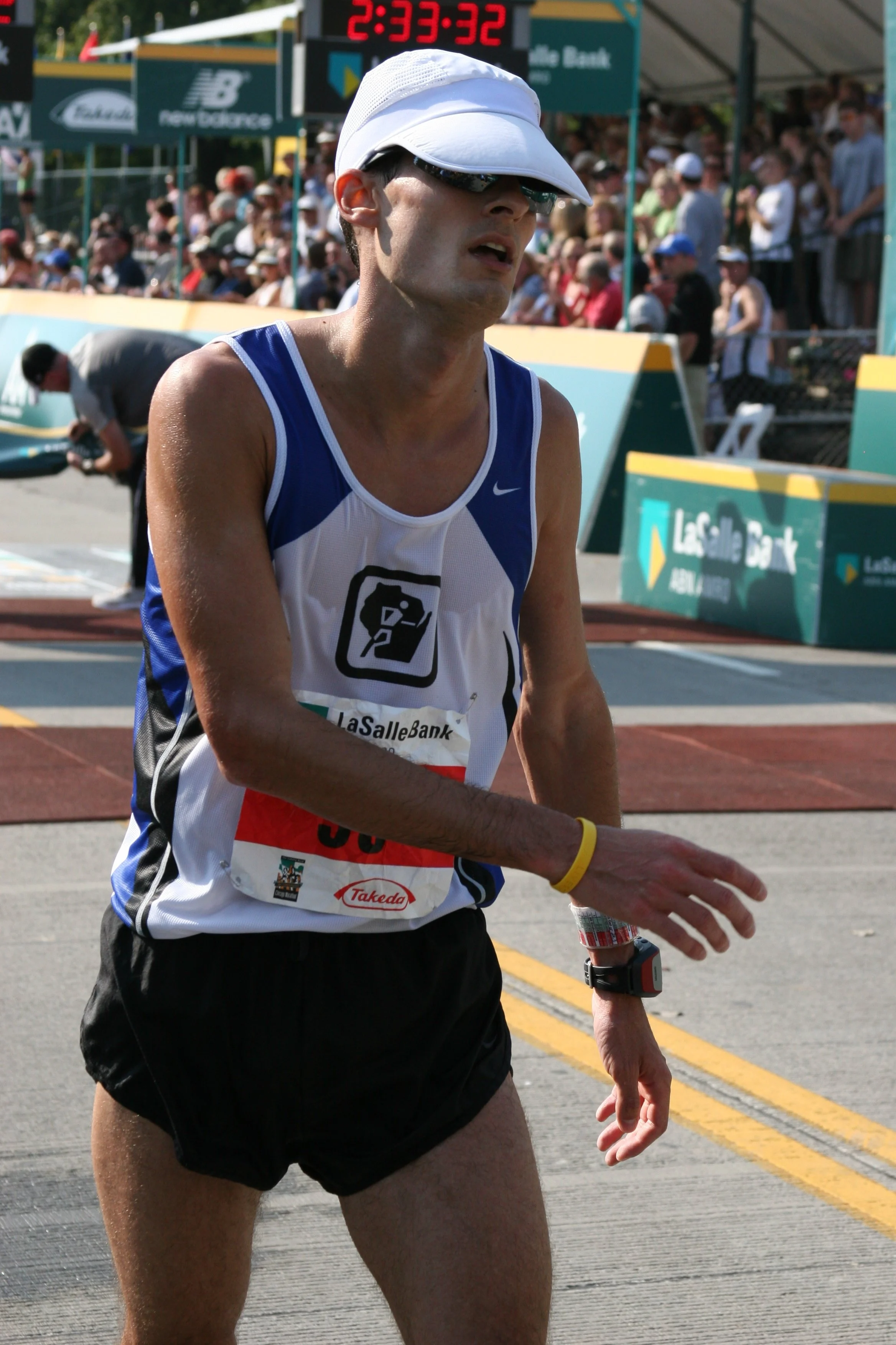 A male marathon runner in athletic attire, including a white cap, sunglasses, and a racing bib, runs near a crowd and a digital clock display at a race event.