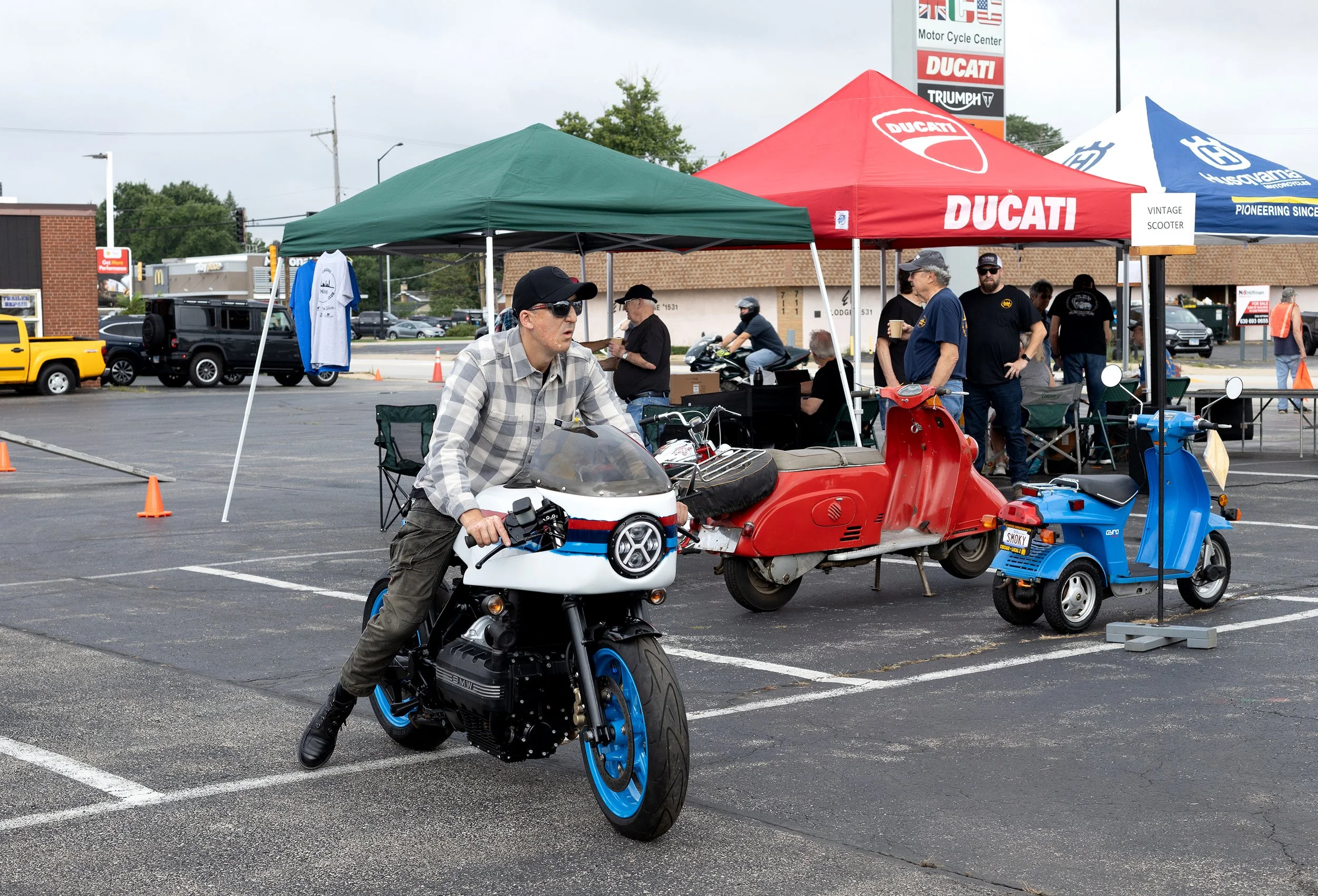 People at a motorcycle event with tents labeled Ducati, vintage scooter, and Husqvarna, showing motorcycles, scooters, and event activities in a parking lot.