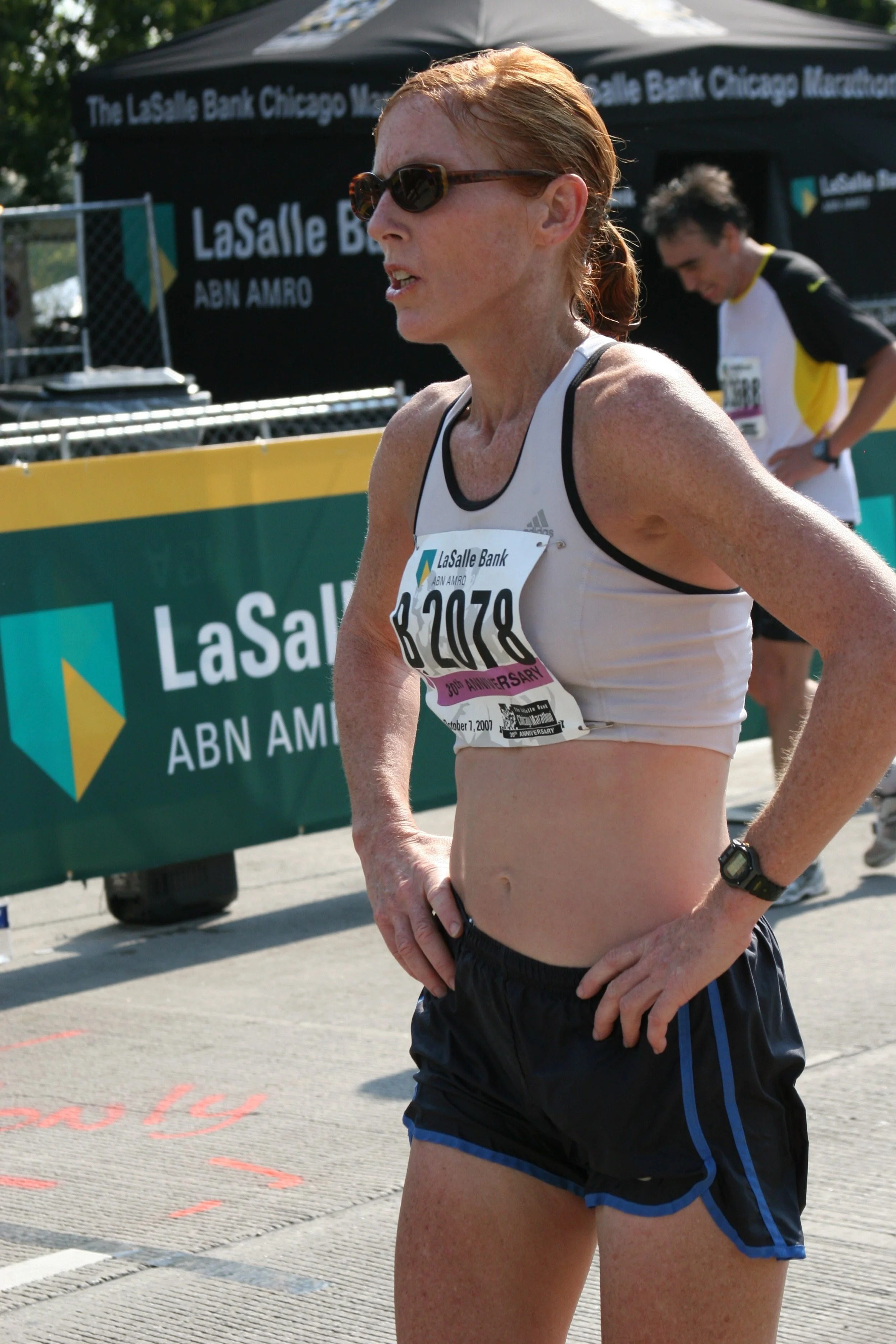 A female runner with red hair, wearing sunglasses, a white running tank top with a bib number 2070, black and blue running shorts, and a black wristwatch, standing during a race next to a green and yellow banner with the text 'LaSalle Bank Chicago Ma