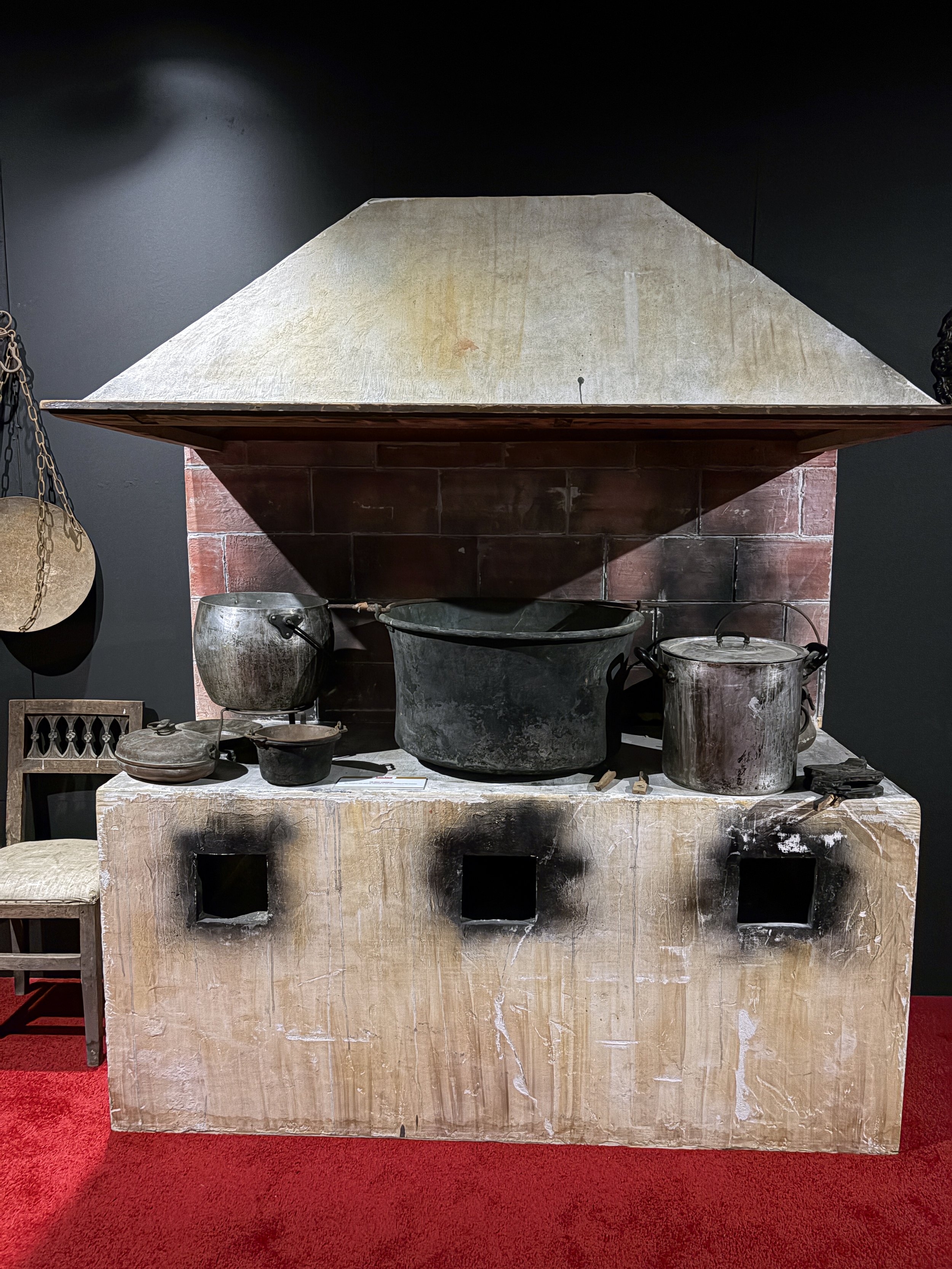 Ancient altar with large stone and metal cooking vessels on top, framed by dark background and red carpet.