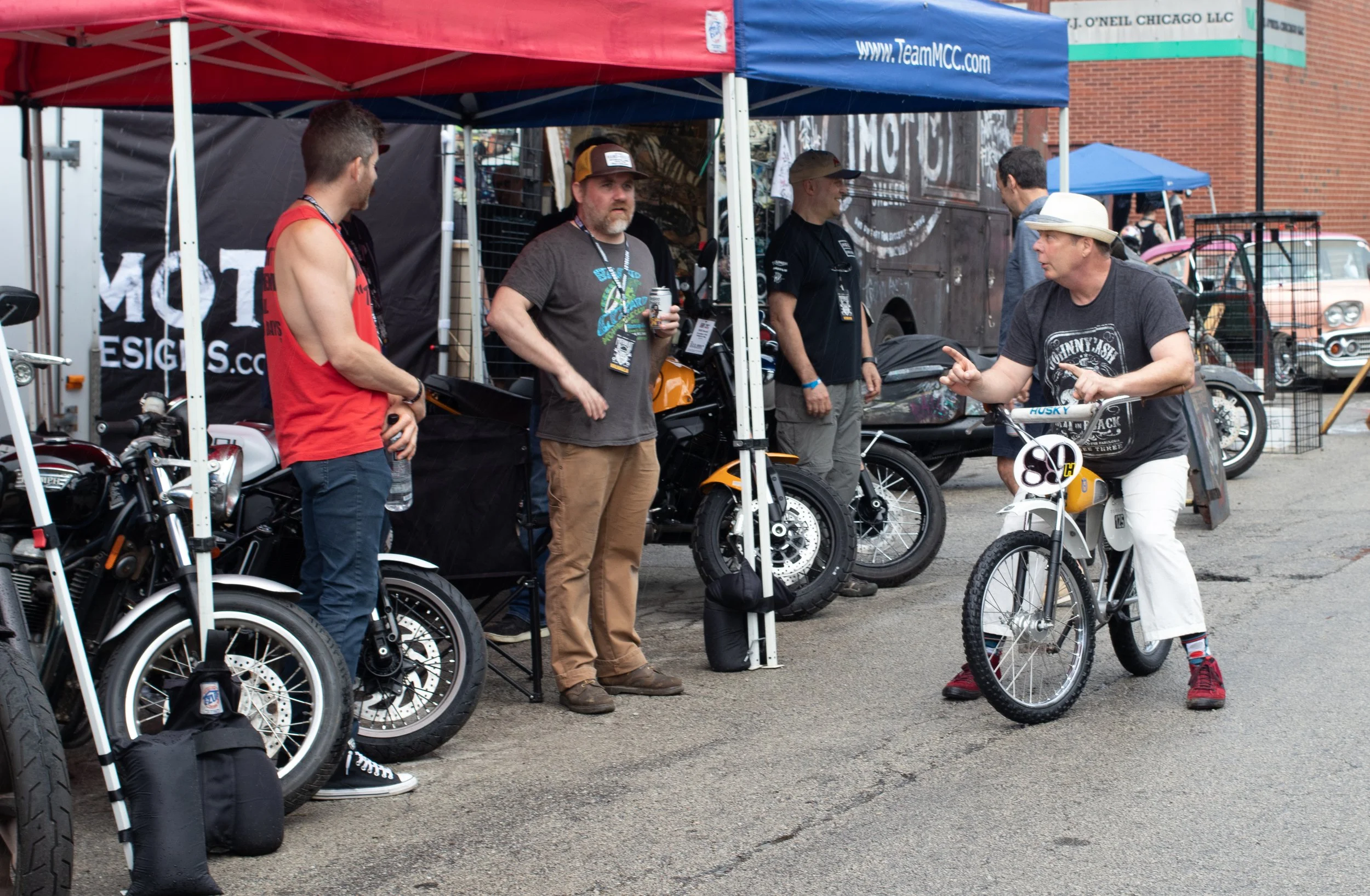 Group of people at an outdoor event with motorcycles and bicycles, enclosed by tents, engaged in conversation.