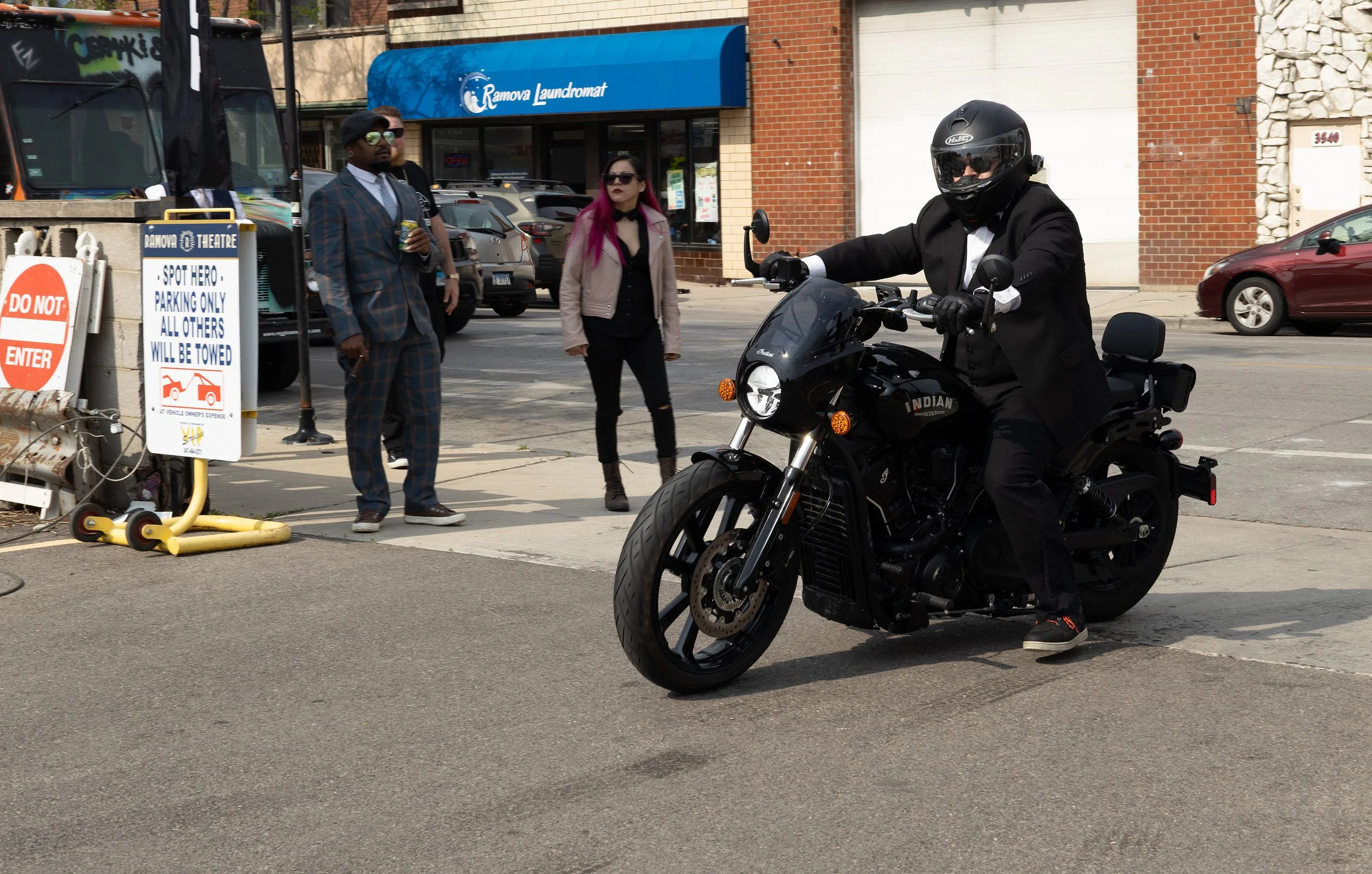 A man in a black suit and helmet riding a black motorcycle on the street. Three people are standing on the sidewalk nearby, two women and one man, watching the scene. There is a blue sign for Ramova Laundromat and some parked cars along the brick bui