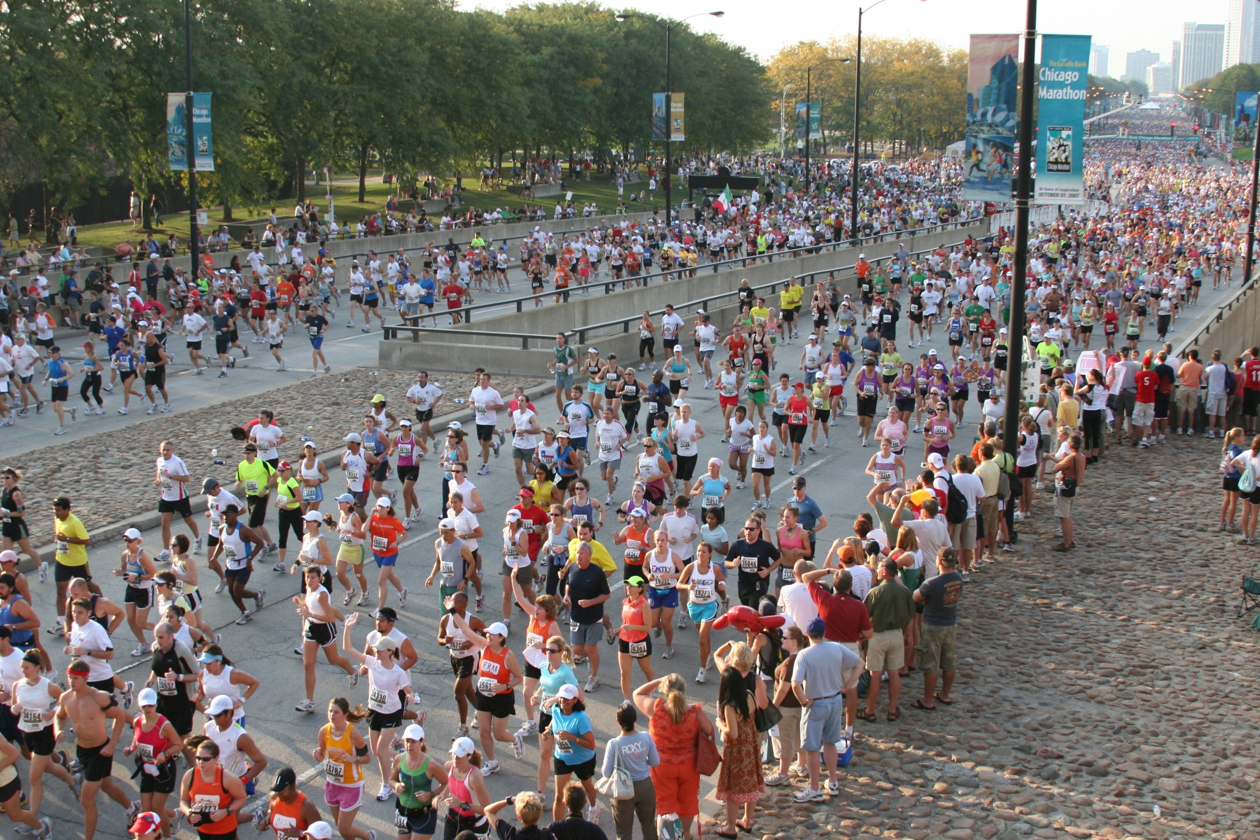 A large crowd participating in a marathon in Chicago, running along a city street lined with trees and spectators, with banners and city buildings in the background.
