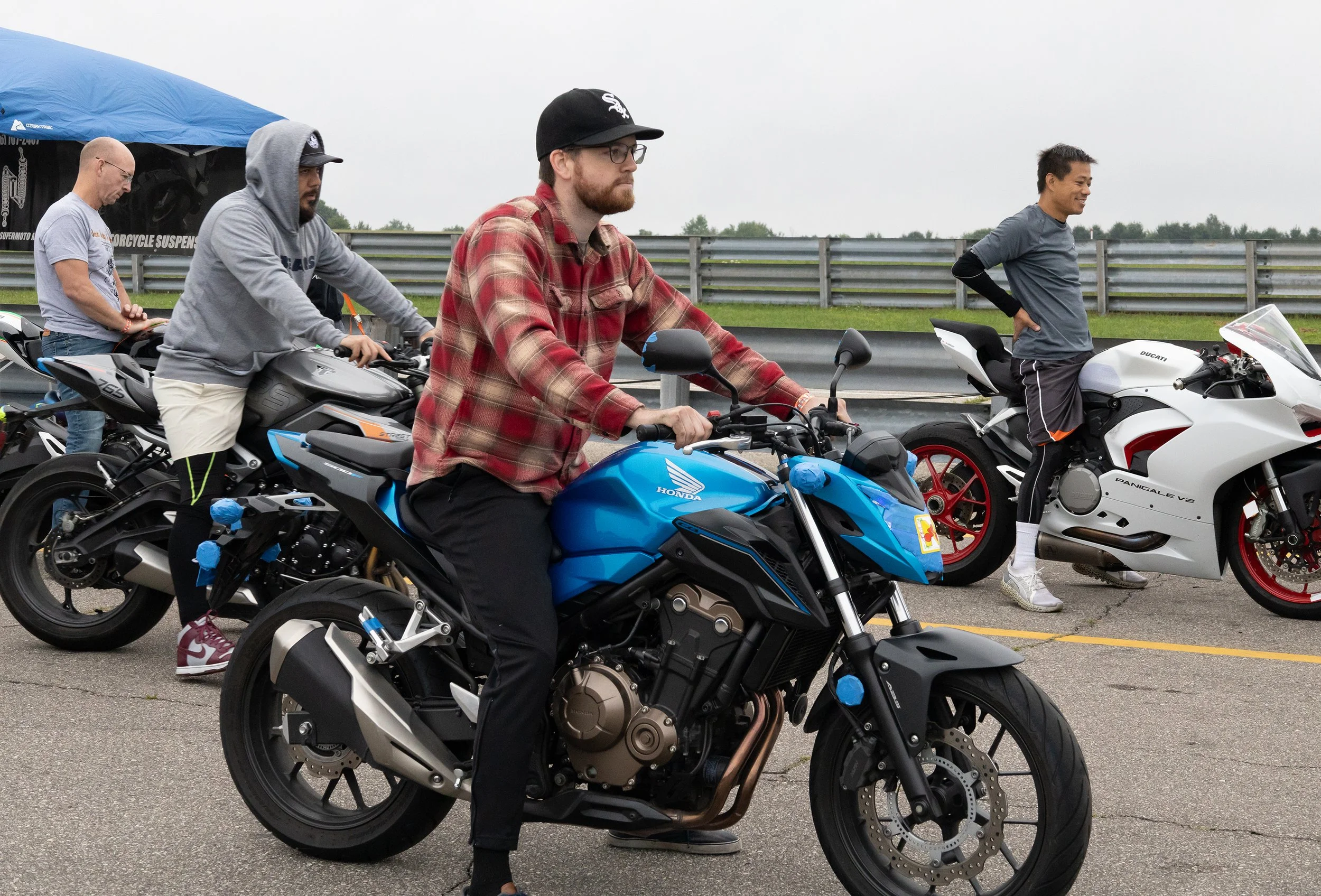 Four men with motorcycles at a track day, one sitting on a blue Honda motorcycle and others near their bikes, with various motorcycle brands and a person in the background under a blue canopy.