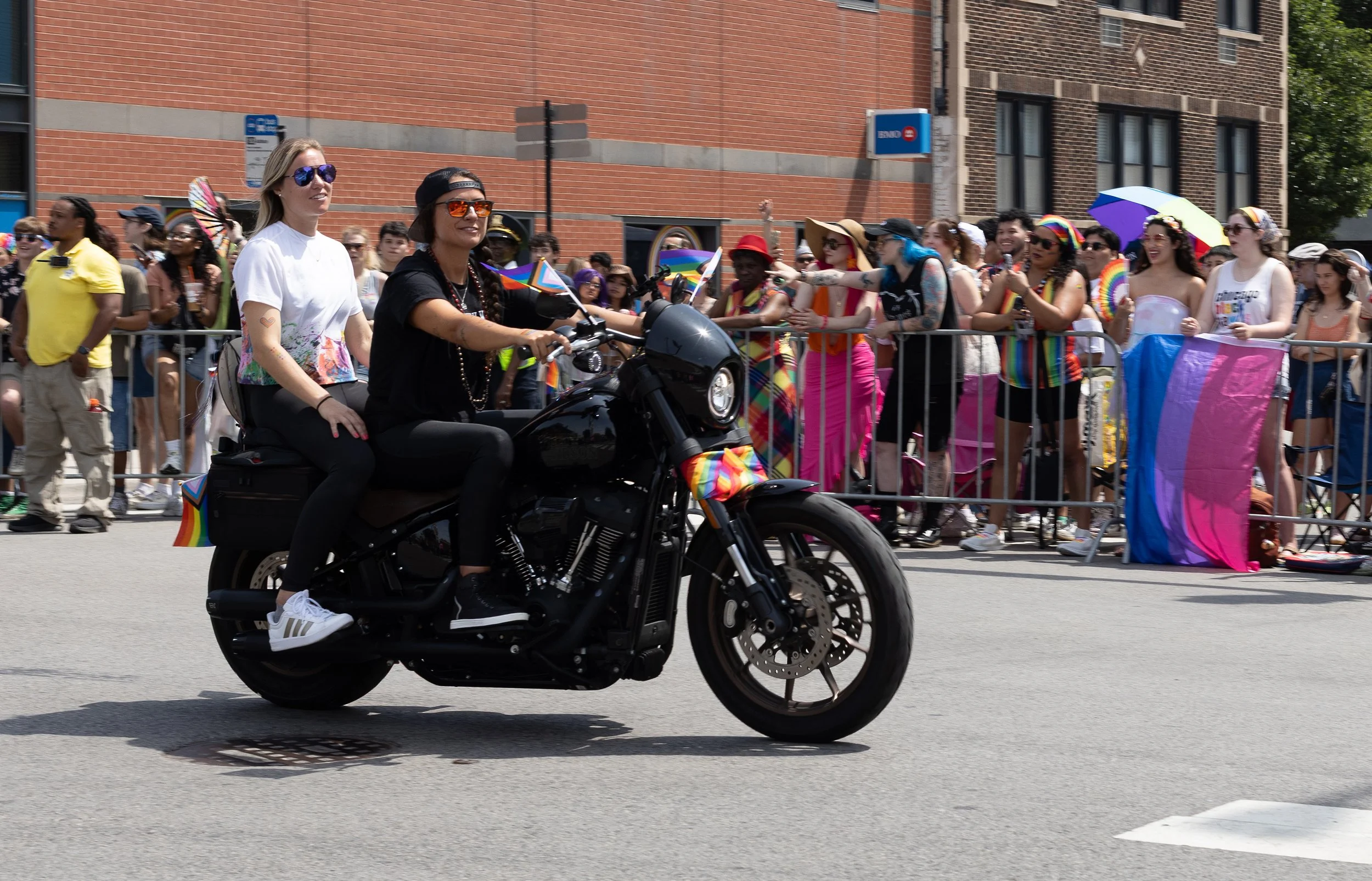 A woman wearing sunglasses, a white t-shirt, and black pants riding a black motorcycle through a parade route with onlookers behind barriers, some waving rainbow flags, wearing colorful clothing, and holding umbrellas that display rainbow colors. The