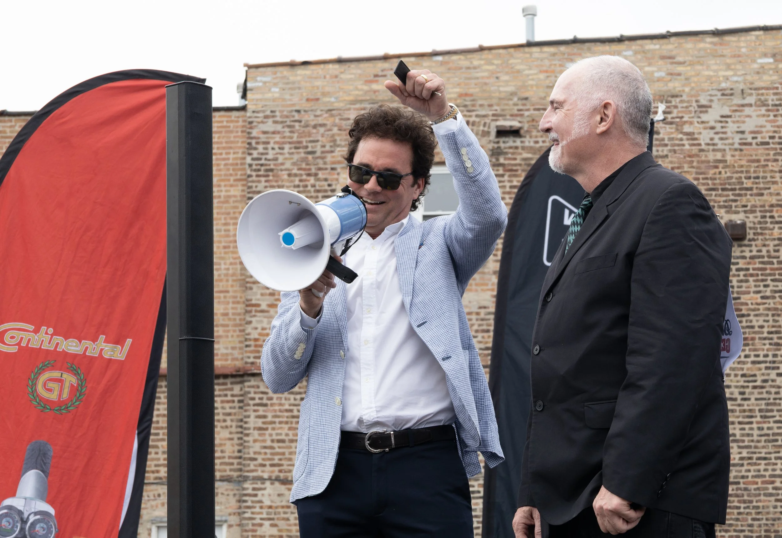 Two men, one with sunglasses and holding a megaphone, shake hands at an outdoor event in front of a brick building, with a red and black banner nearby.