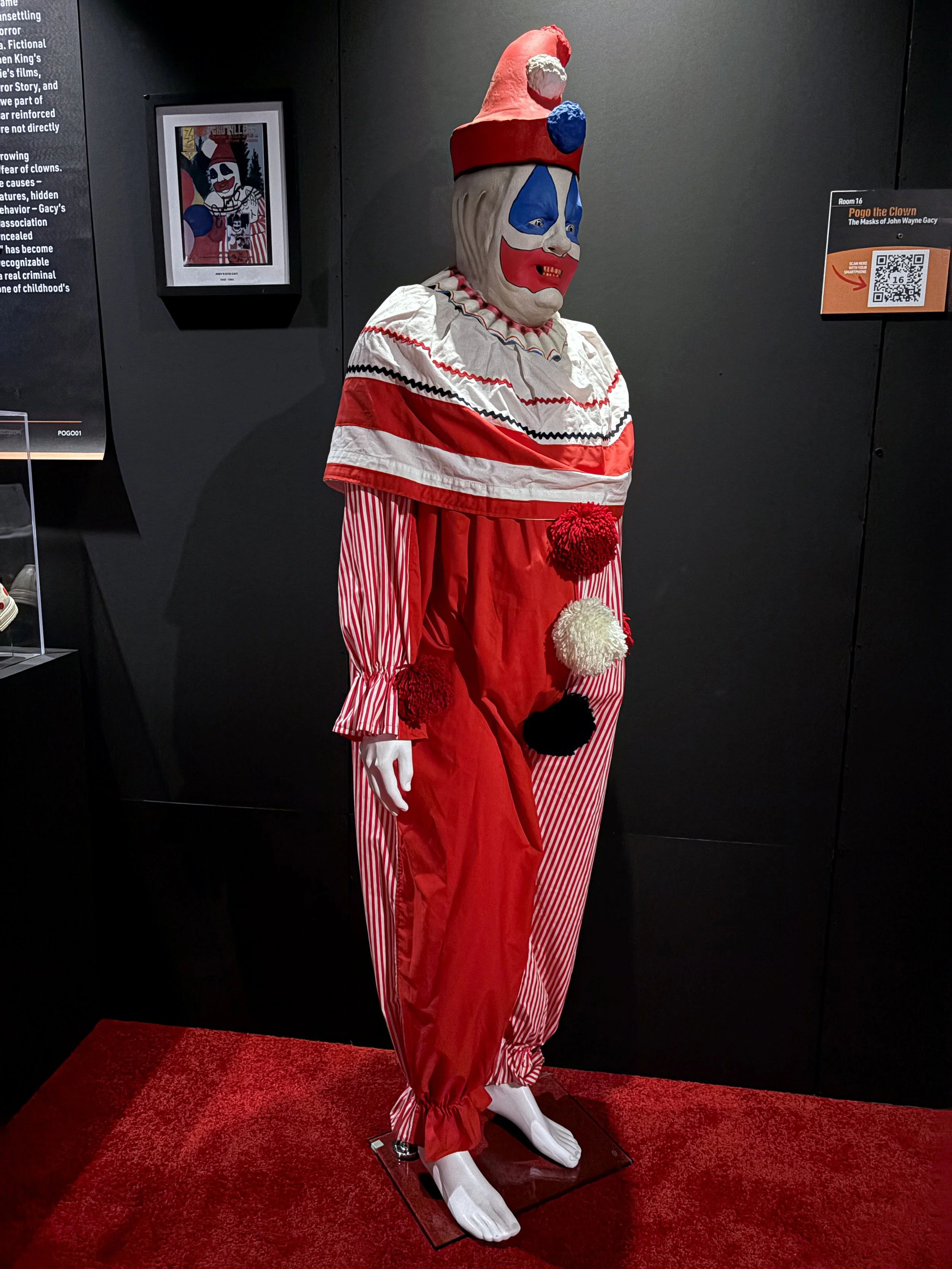 A vintage clown costume mannequin dressed in a red and white striped jumpsuit with large pom-poms and a ruffled collar, wearing a white clown mask with blue, red, and white painted features and a red, pink, and blue hat, displayed on a red carpet in 