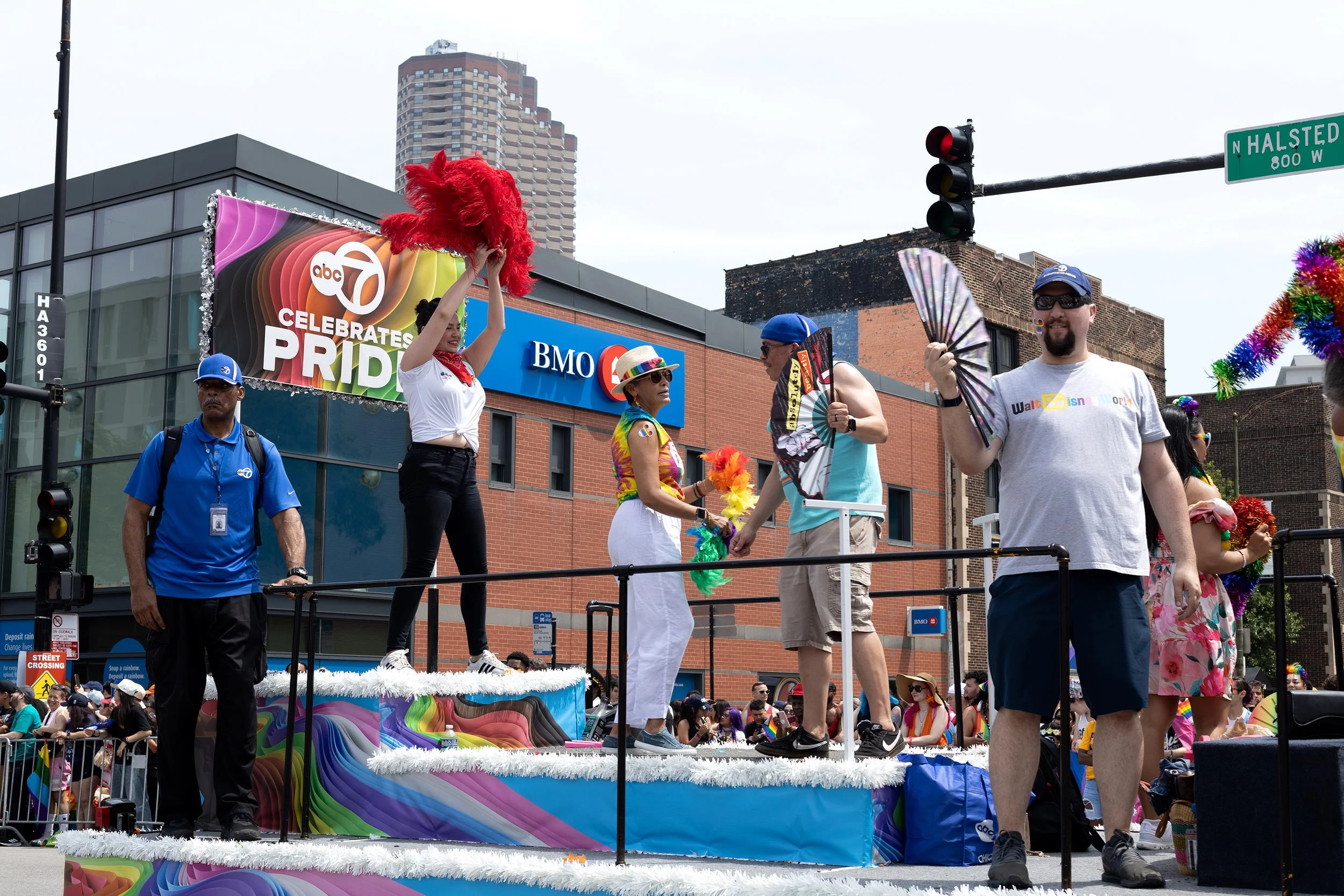 People participating in a Pride parade, standing on a decorated float with rainbow-colored designs. Some are wearing rainbow accessories, and one person is holding a rainbow fan. A large sign reads 'ABC 7 Celebrates Pride' in the background.
